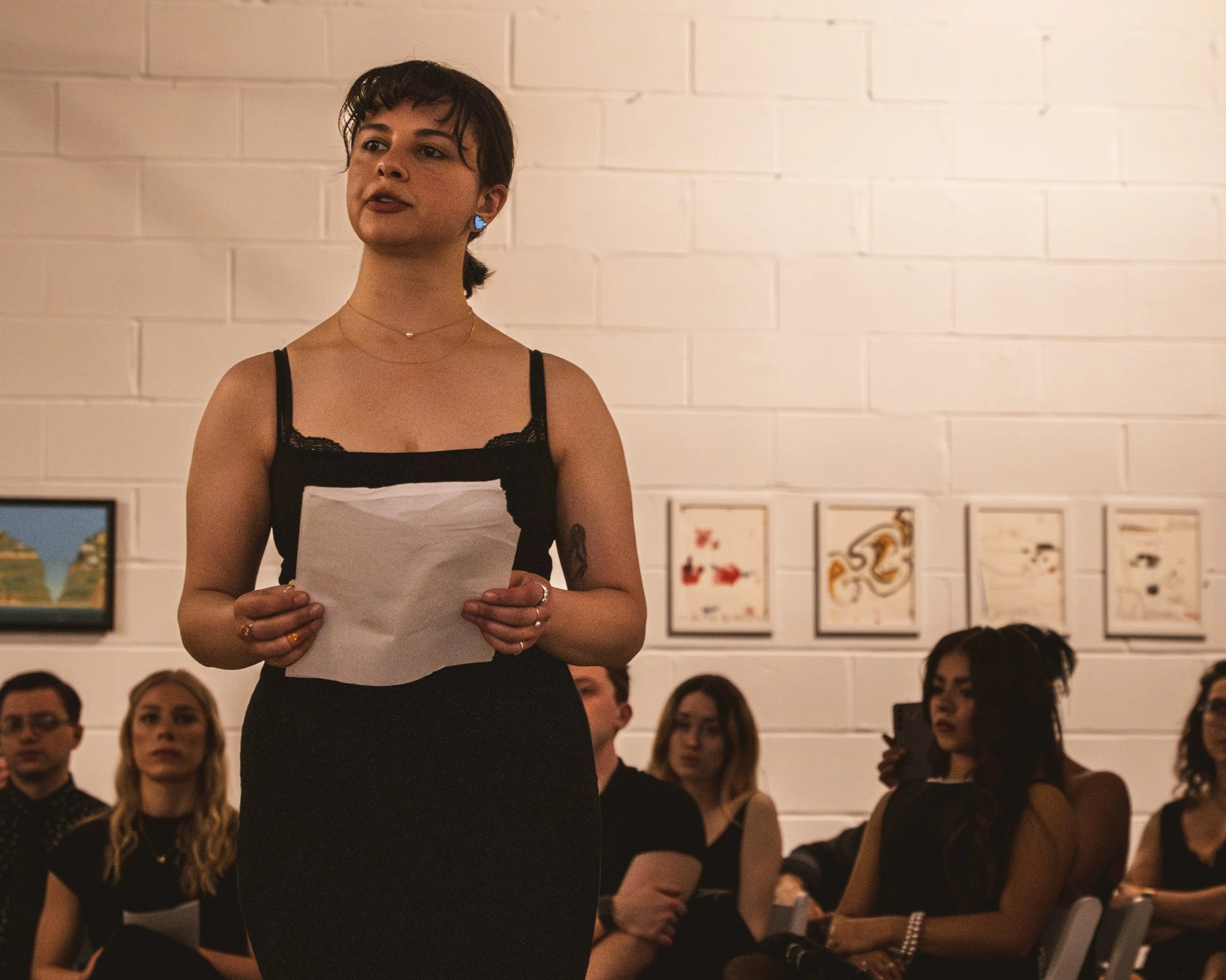 A woman with short dark hair, wearing a black spaghetti strap top, stands and speaks to an audience in an art gallery, holding papers in her hands. The background shows a white brick wall with framed artwork and several seated listeners, including a woman with long curly hair and a woman with long dark hair and glasses.