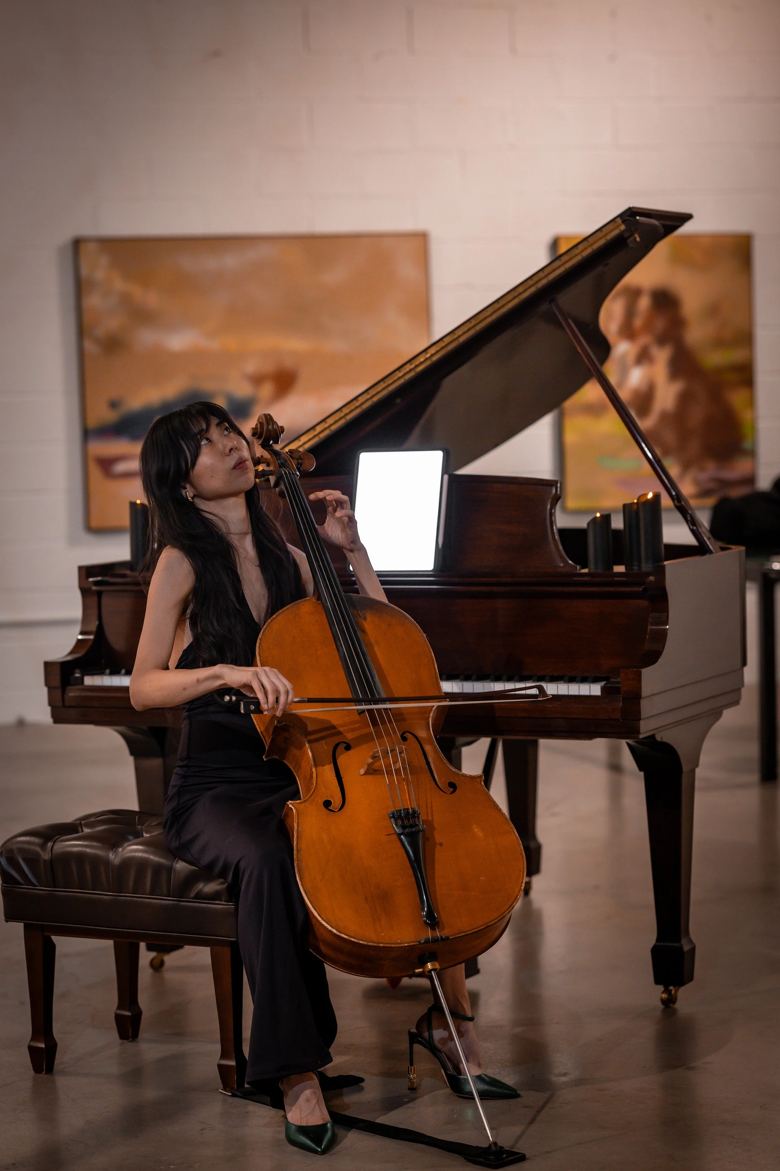 A woman playing the cello while sitting on a leather bench, with a grand piano behind her in an art gallery setting.