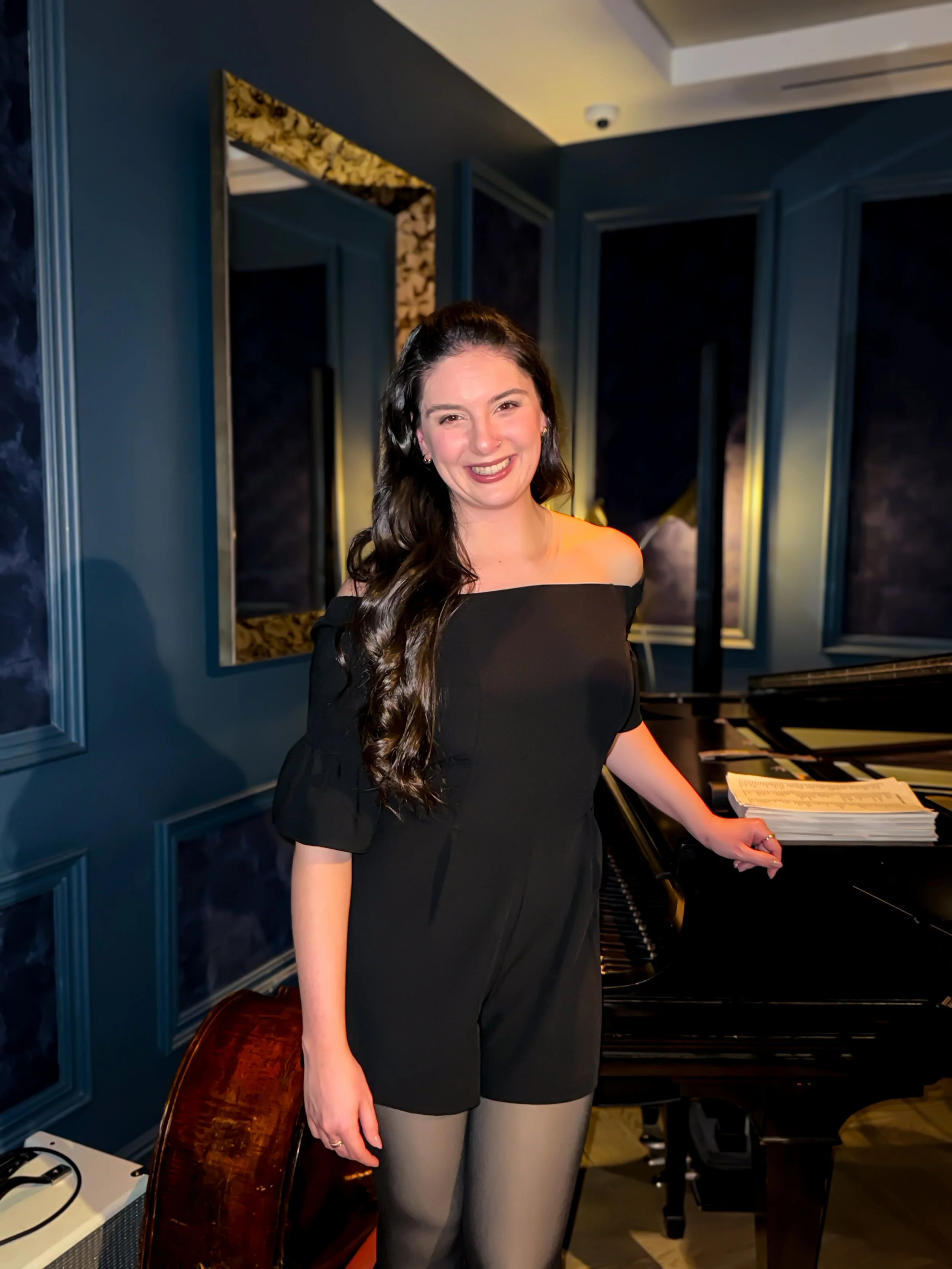 A smiling woman with long dark hair wearing a black off-the-shoulder dress and beige tights, standing next to a grand piano in a room with dark blue walls and gold-framed mirrors.