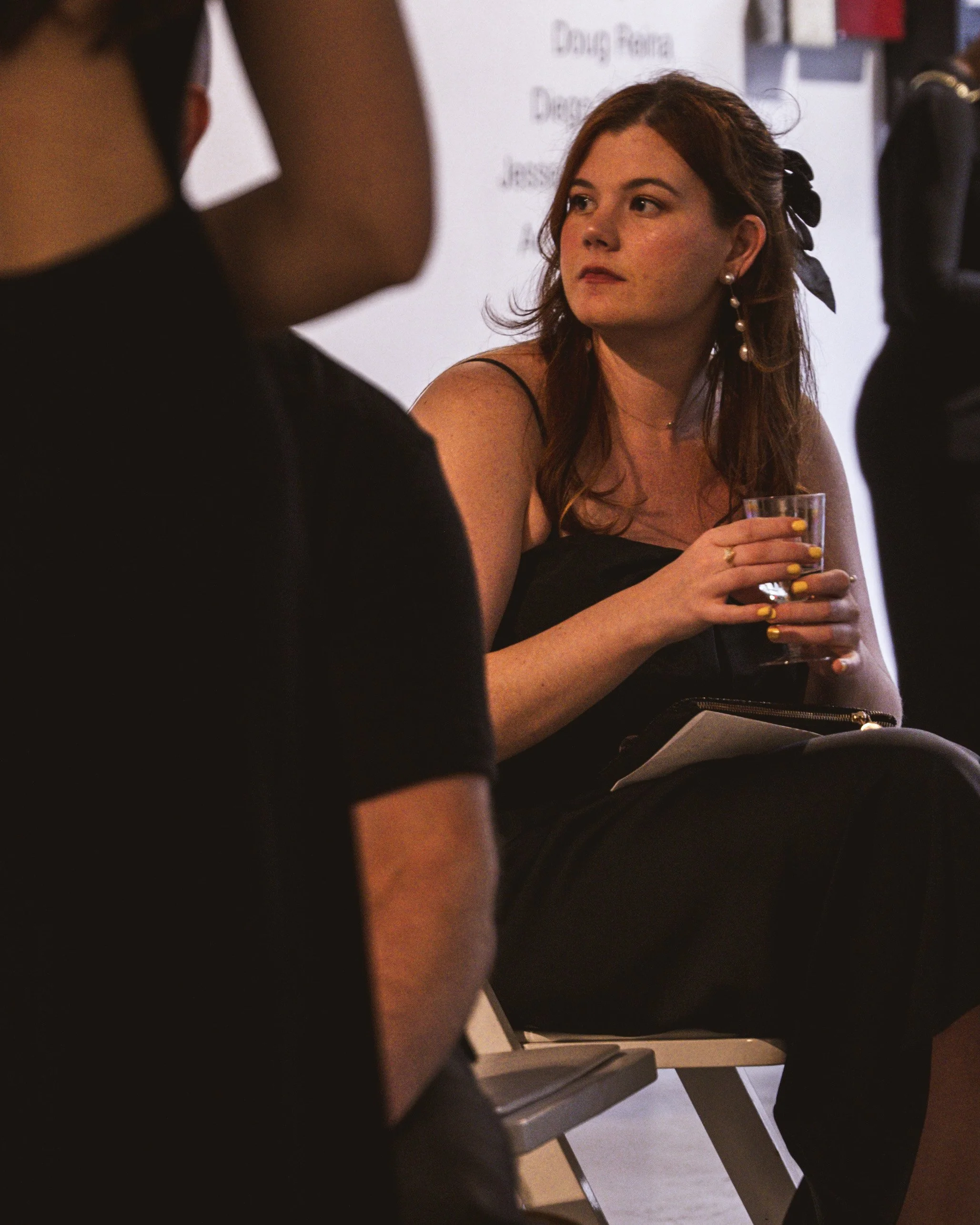 A woman with red hair, wearing a black dress and dangling pearl earrings, sitting and holding a glass, appears to be listening or waiting at an indoor event.