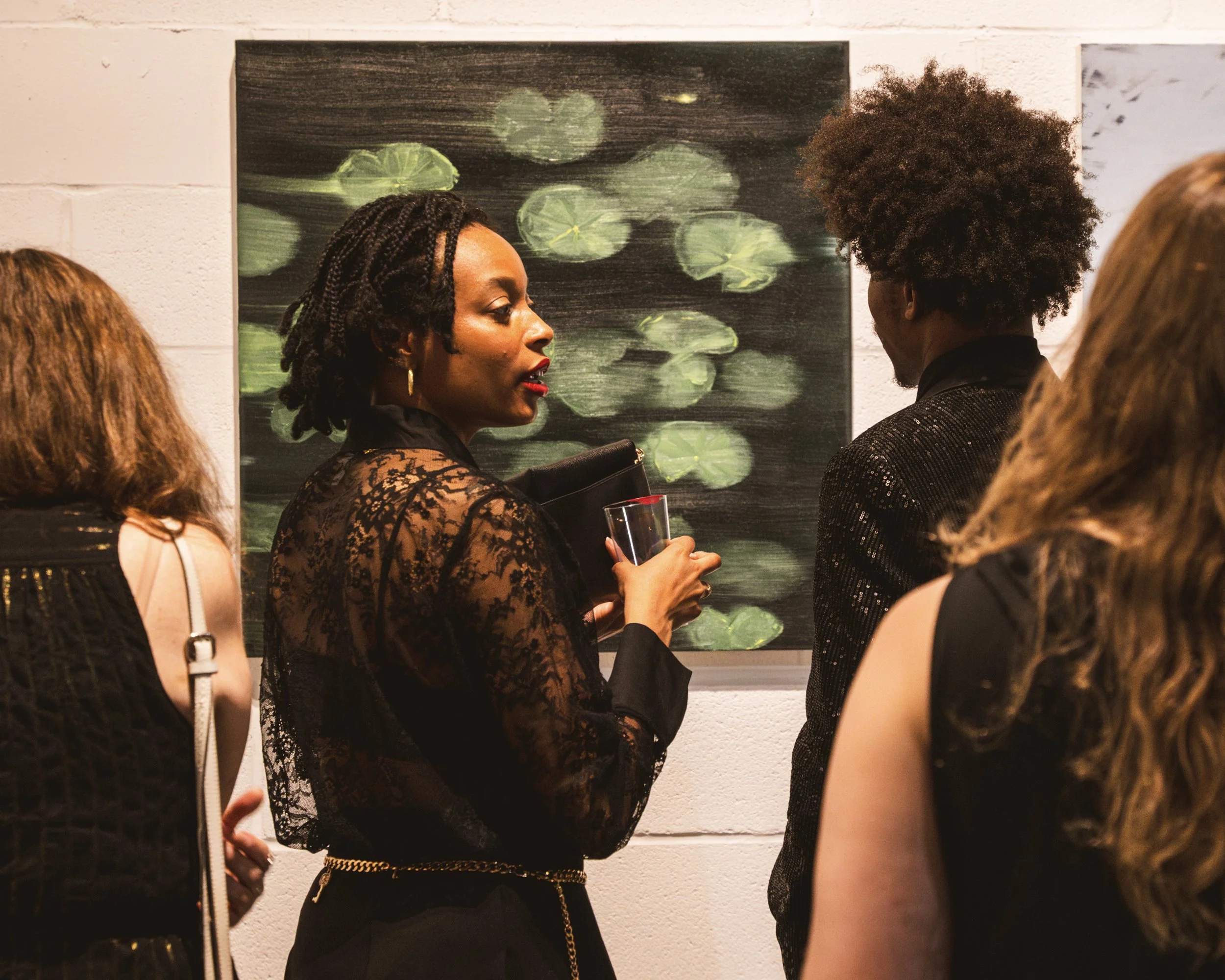Group of women at an art gallery, engaging in conversation, with a woman in a lace top holding a glass, standing in front of an abstract painting with green and black hues.