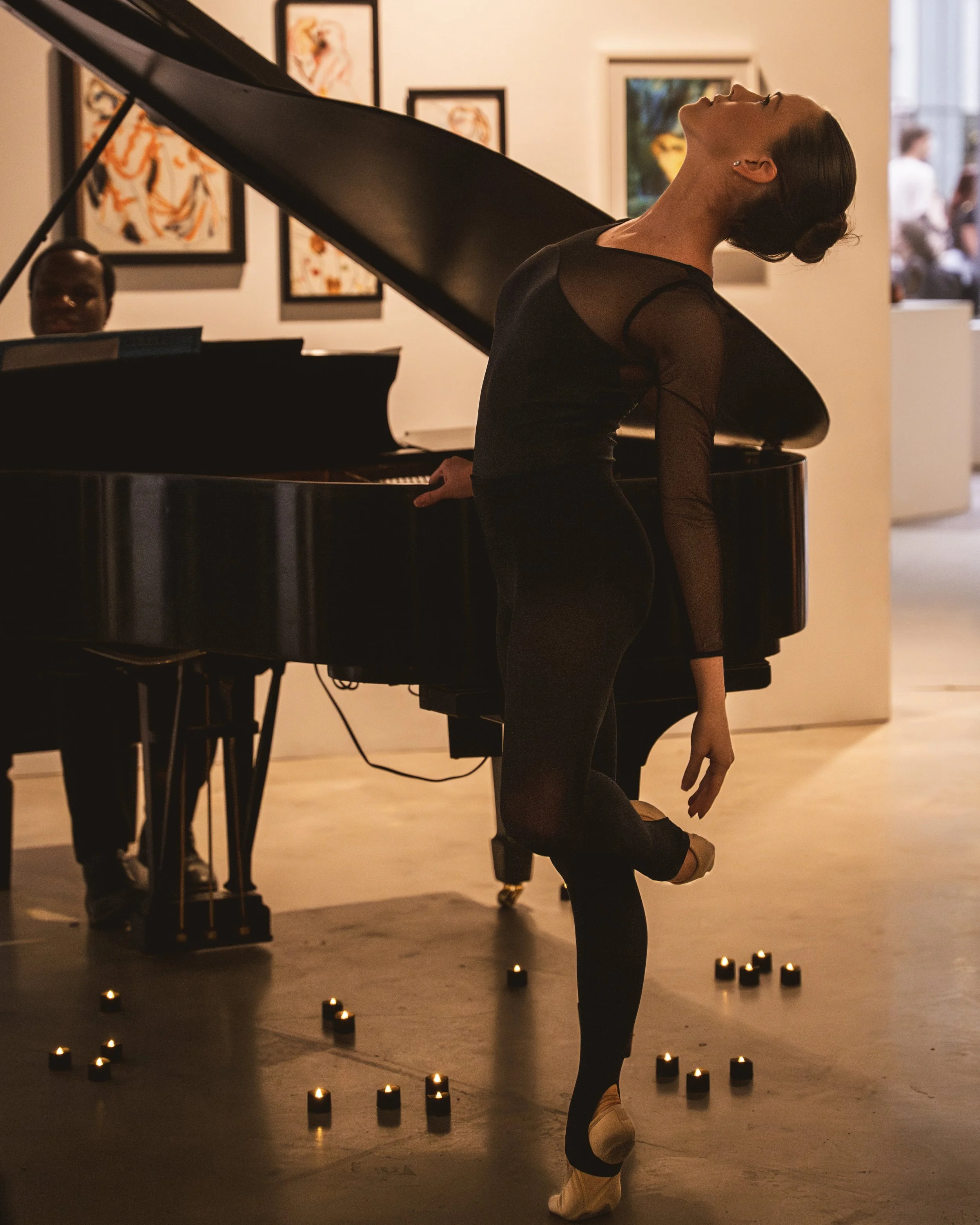 A woman in a black, sheer, long-sleeved dance or ballet costume leaning against a grand piano with a person playing it. The scene is set in an art gallery or similar space with artworks on the wall and small candles on the floor.