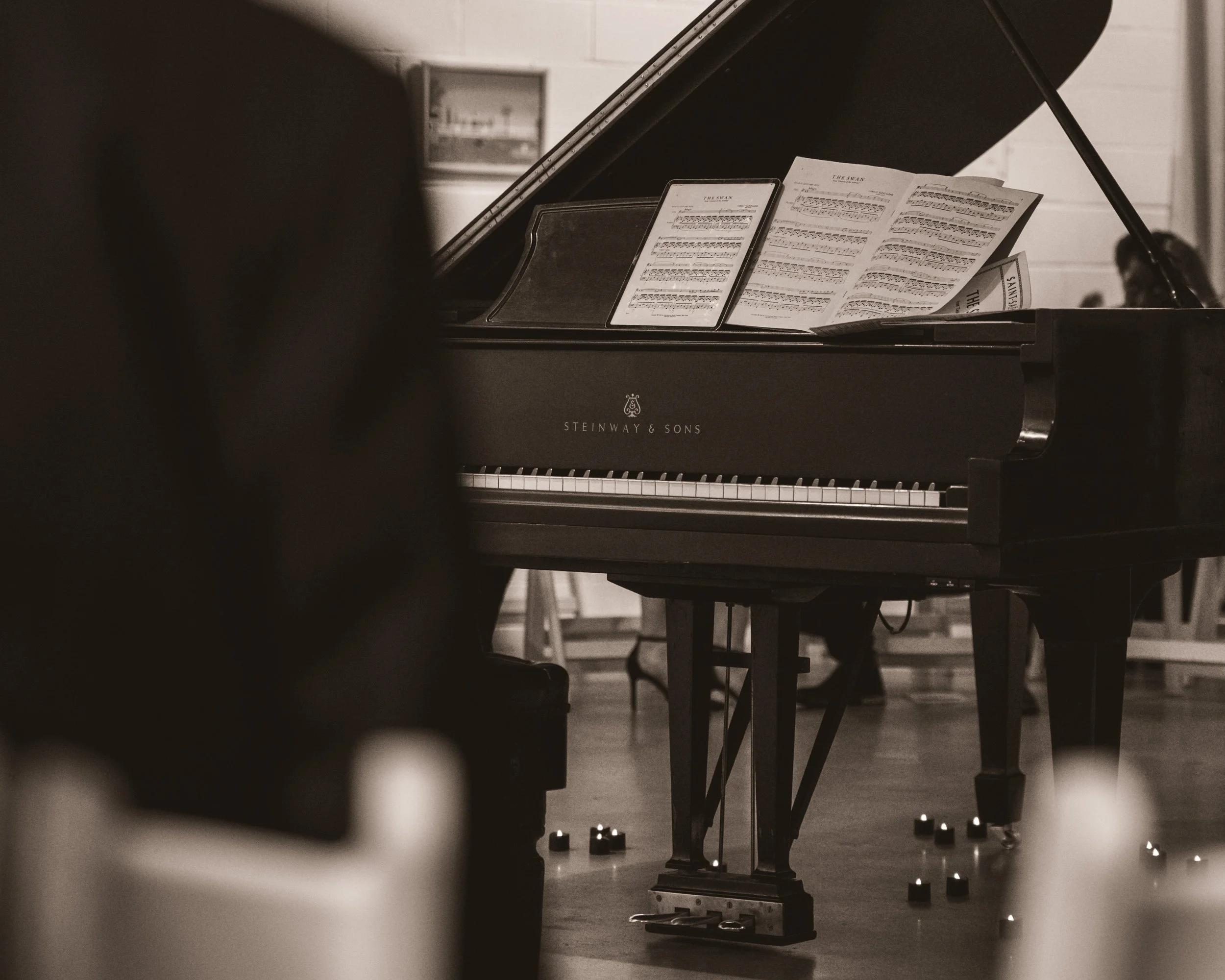 A grand piano with music sheets on music stands inside a room, with small candles on the floor in front of it.