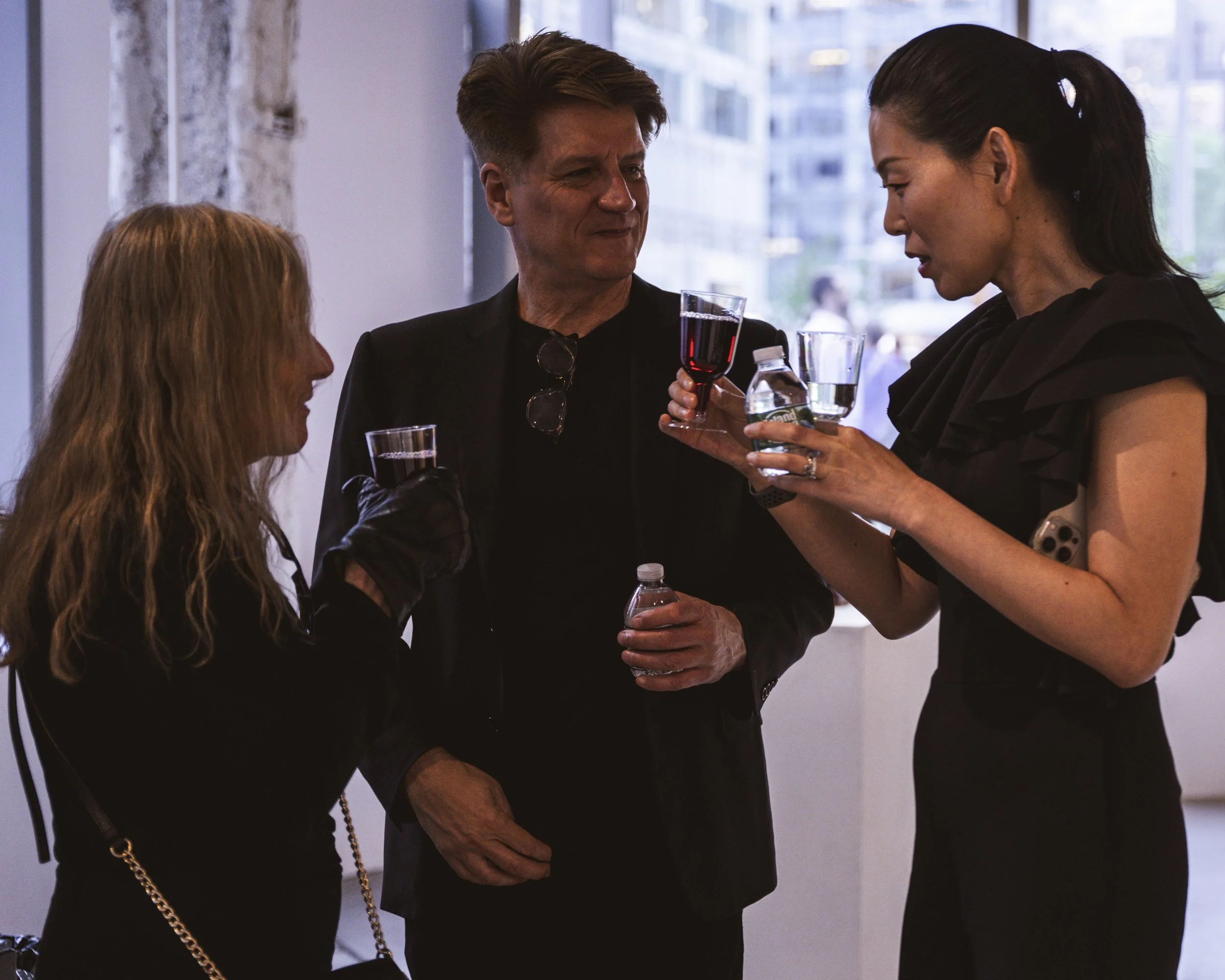 Three people in conversation at an indoor social event, with the woman on the right holding a glass of red wine and a water bottle, and the man in the middle holding a small water bottle.