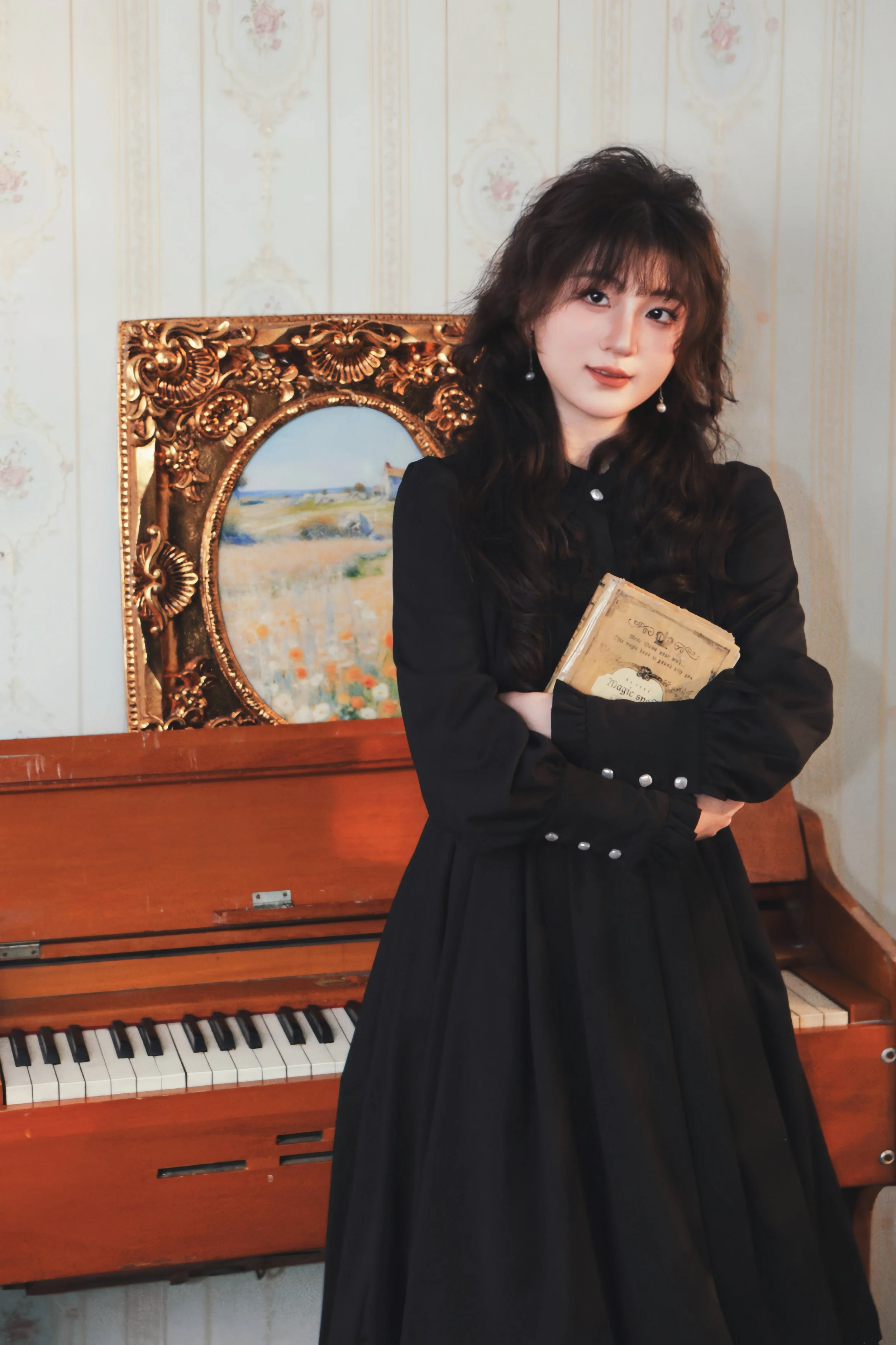 Young woman with long dark hair, wearing a black dress with pearl buttons, holding a vintage book, standing in front of a wooden piano and a framed painting of a landscape.