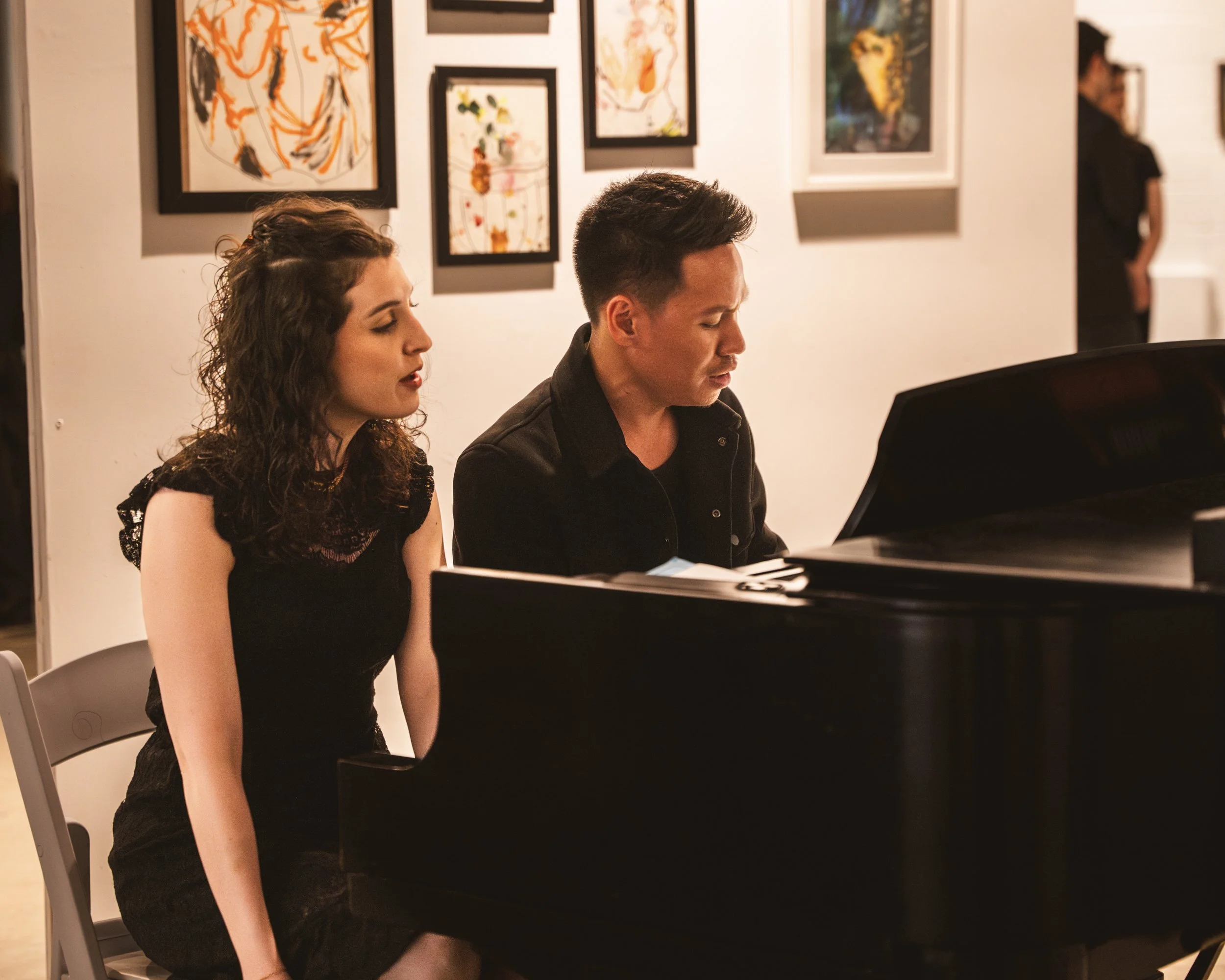 A woman and man sitting at a black grand piano, performing together in an art gallery with colorful abstract paintings on the white walls.