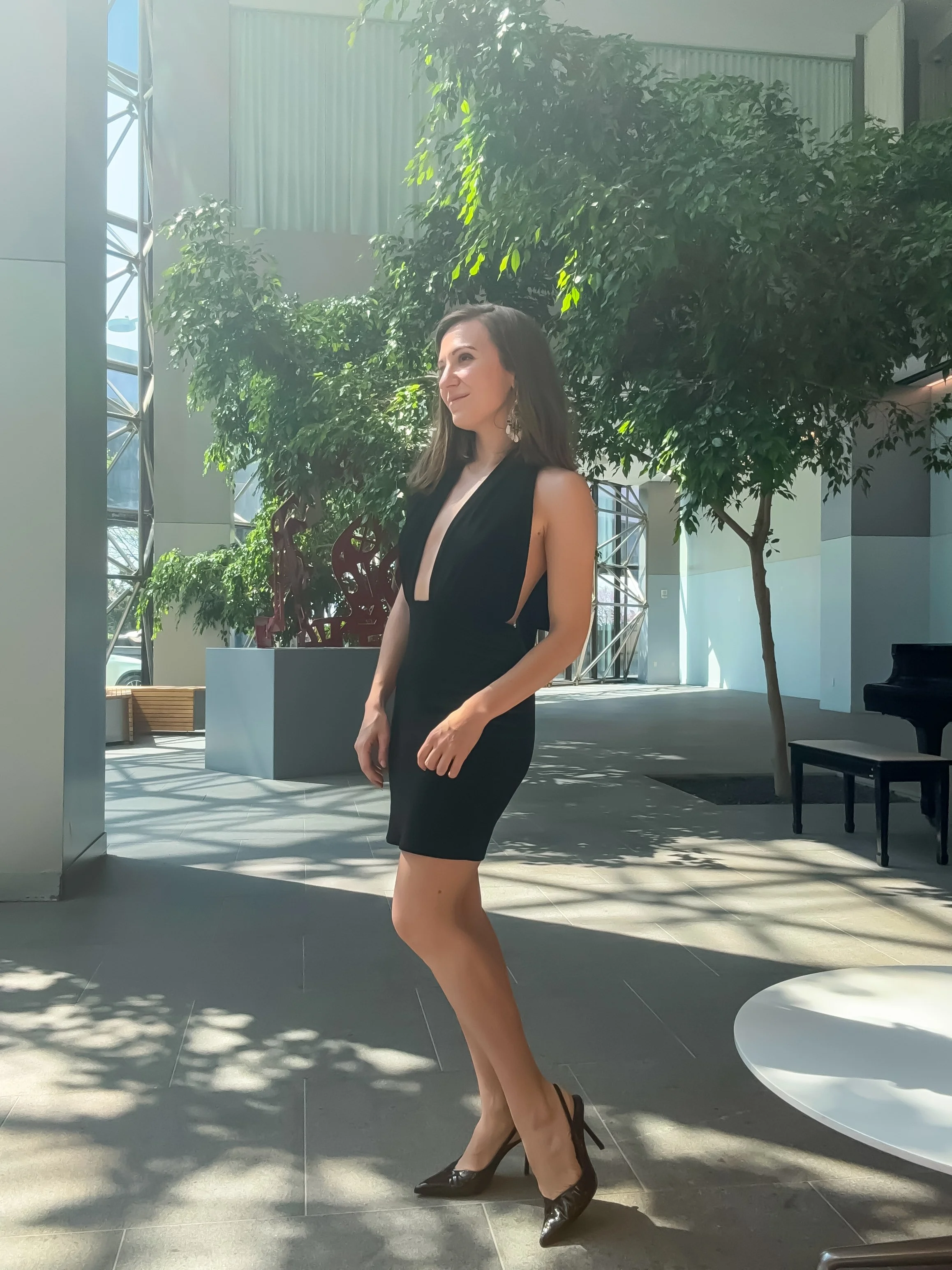 A woman in a black dress and high heels standing indoors near large trees with sunlight streaming in.