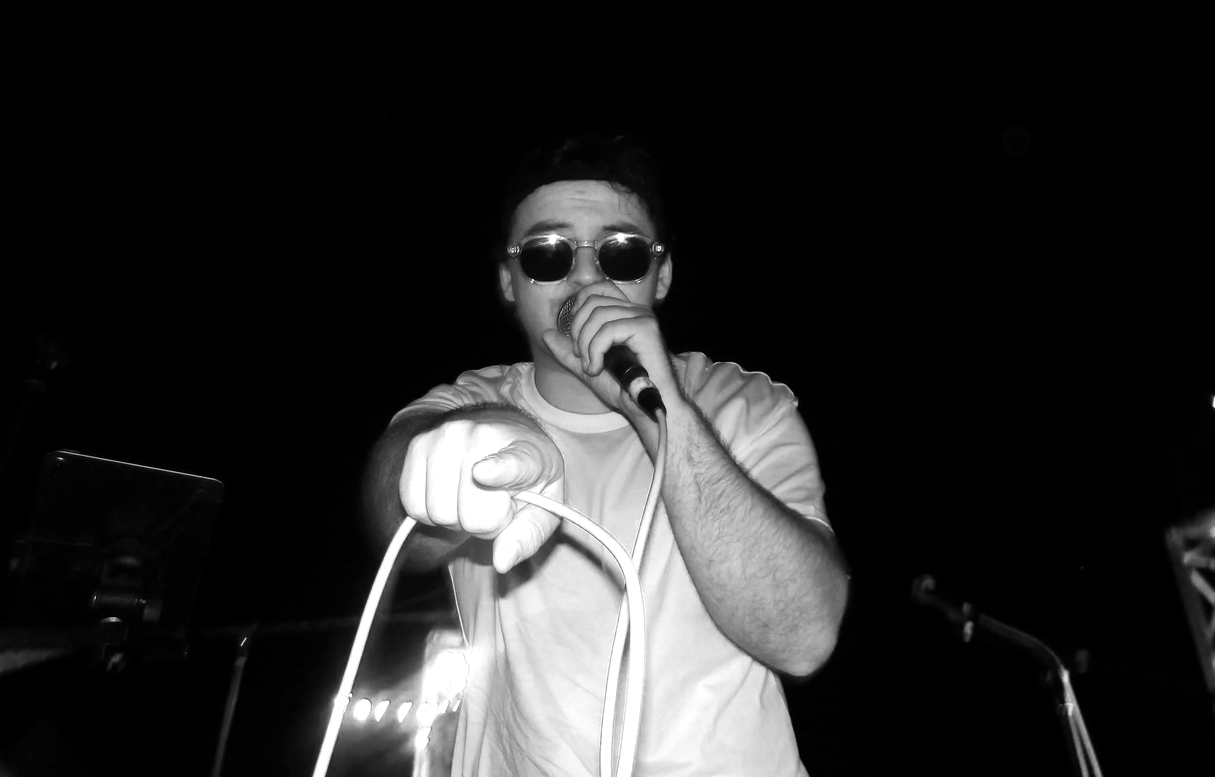 Un homme portant des lunettes de soleil, tenant un micro et pointant vers l'avant. La photo est en noir et blanc.