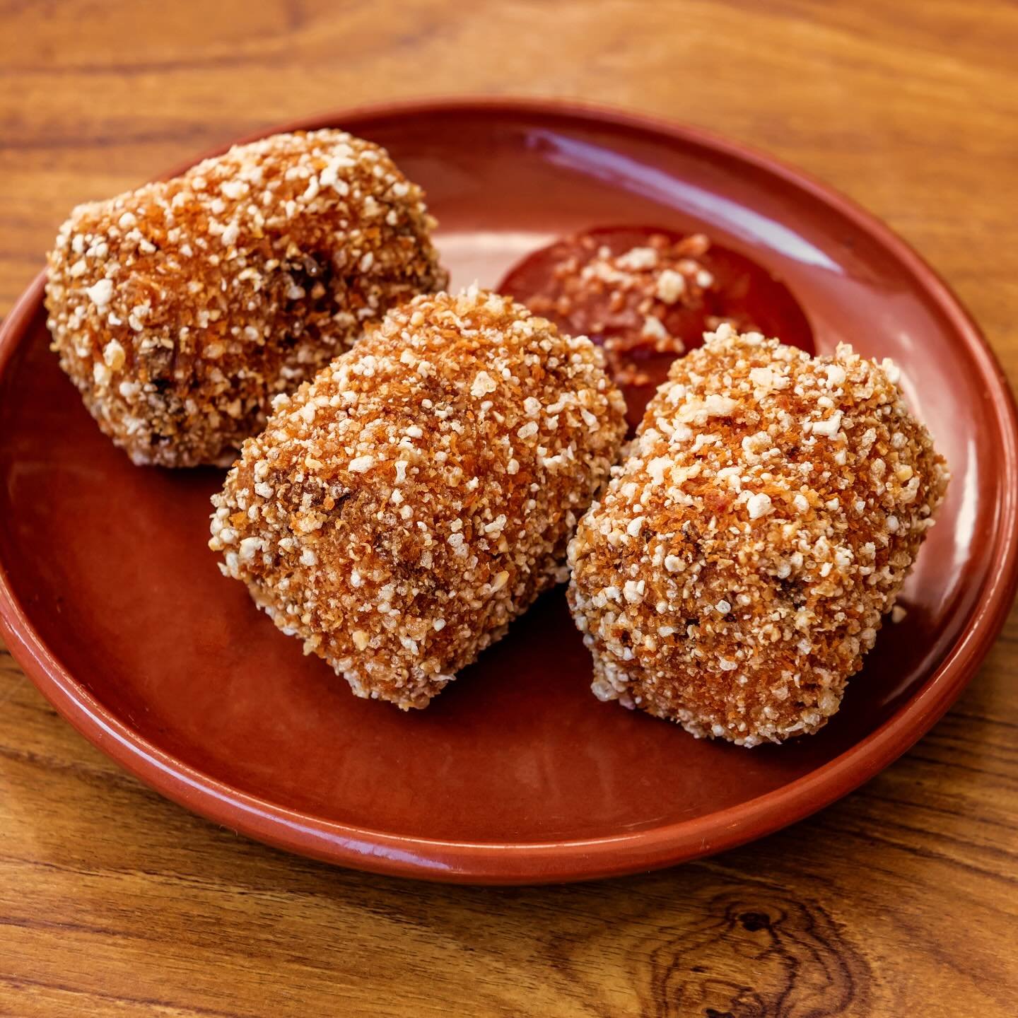 Three breaded and fried food items on a brown plate with a side of dipping sauce, placed on a wooden surface.
