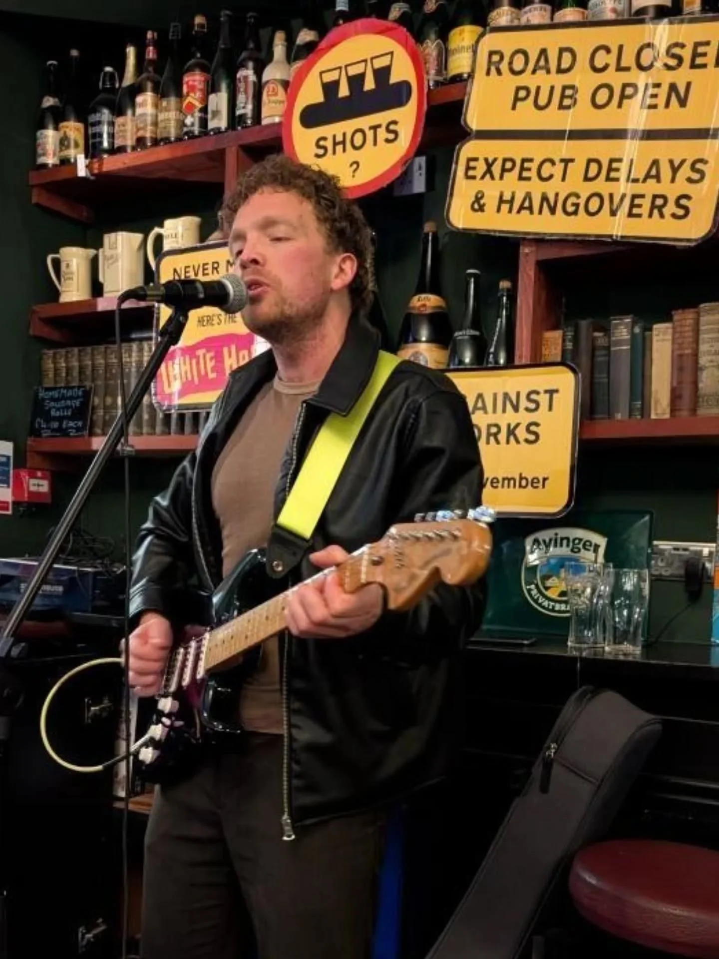 A man playing an electric guitar and singing into a microphone in a pub setting, with various signs and bottles in the background.