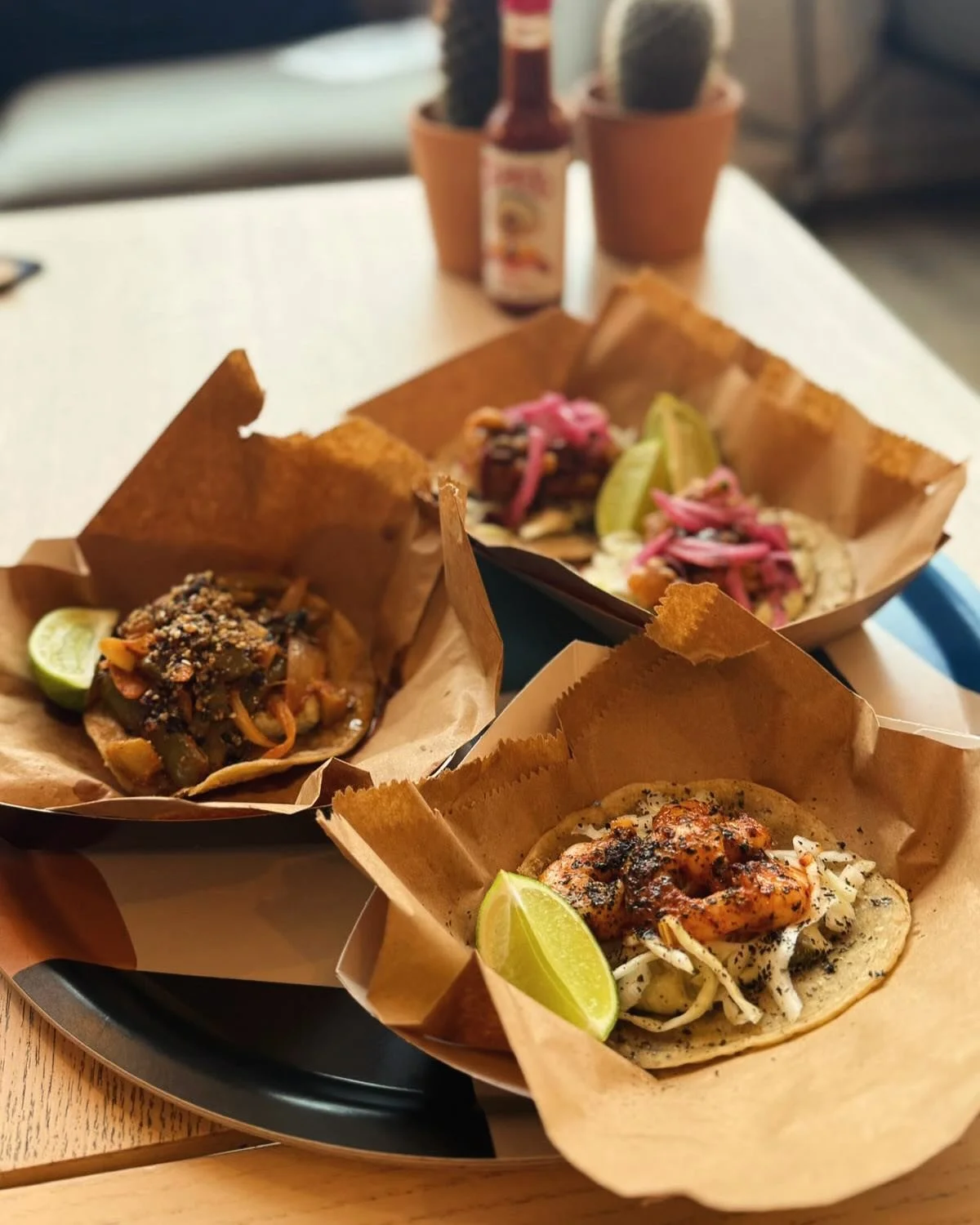 Three tacos with various fillings served on a tray with lime wedges, on a wooden table with potted cacti and a hot sauce bottle in the background.