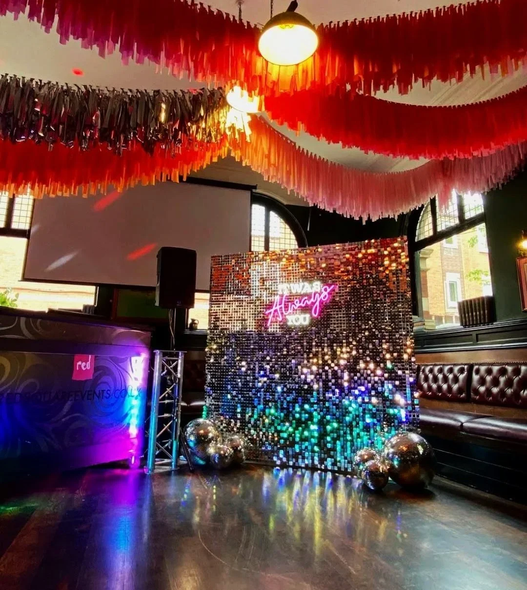 Event setup with a sparkly backdrop that has a pink neon sign reading 'AWAY' and 'Aisage,' metallic balloons, a leather bench, and hanging pink and red paper streamers from the ceiling in a decorated room.