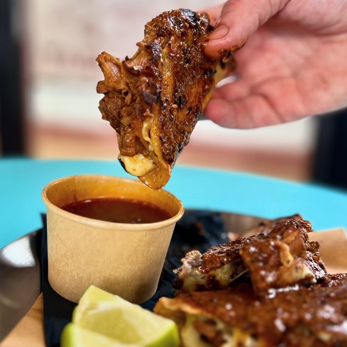 Close-up of a hand dipping a birria taco into a pot of beef consommé, with lime wedges on a plate.