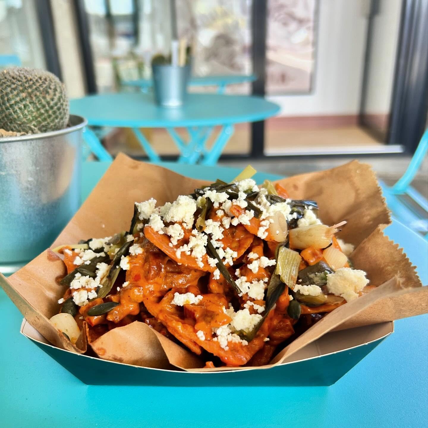 A bowl of Mexican food with vegetables and crumbled cheese on top, served on a brown paper tray, placed on a turquoise table in a bright, modern dining area.