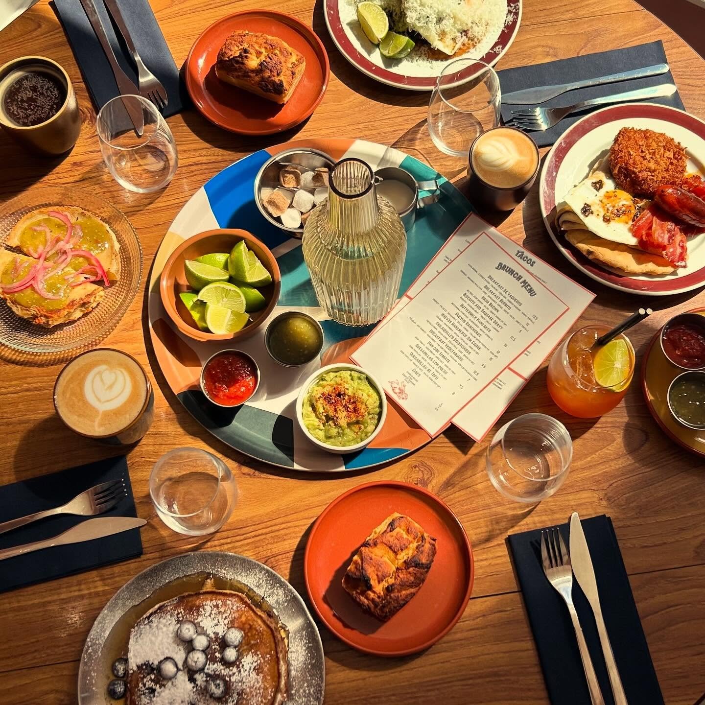 A wooden table set with assorted food and drinks, including tacos, a plate with a burger, lime wedges, a coffee cup, juice glasses, a dessert, and a brunch menu in the center.