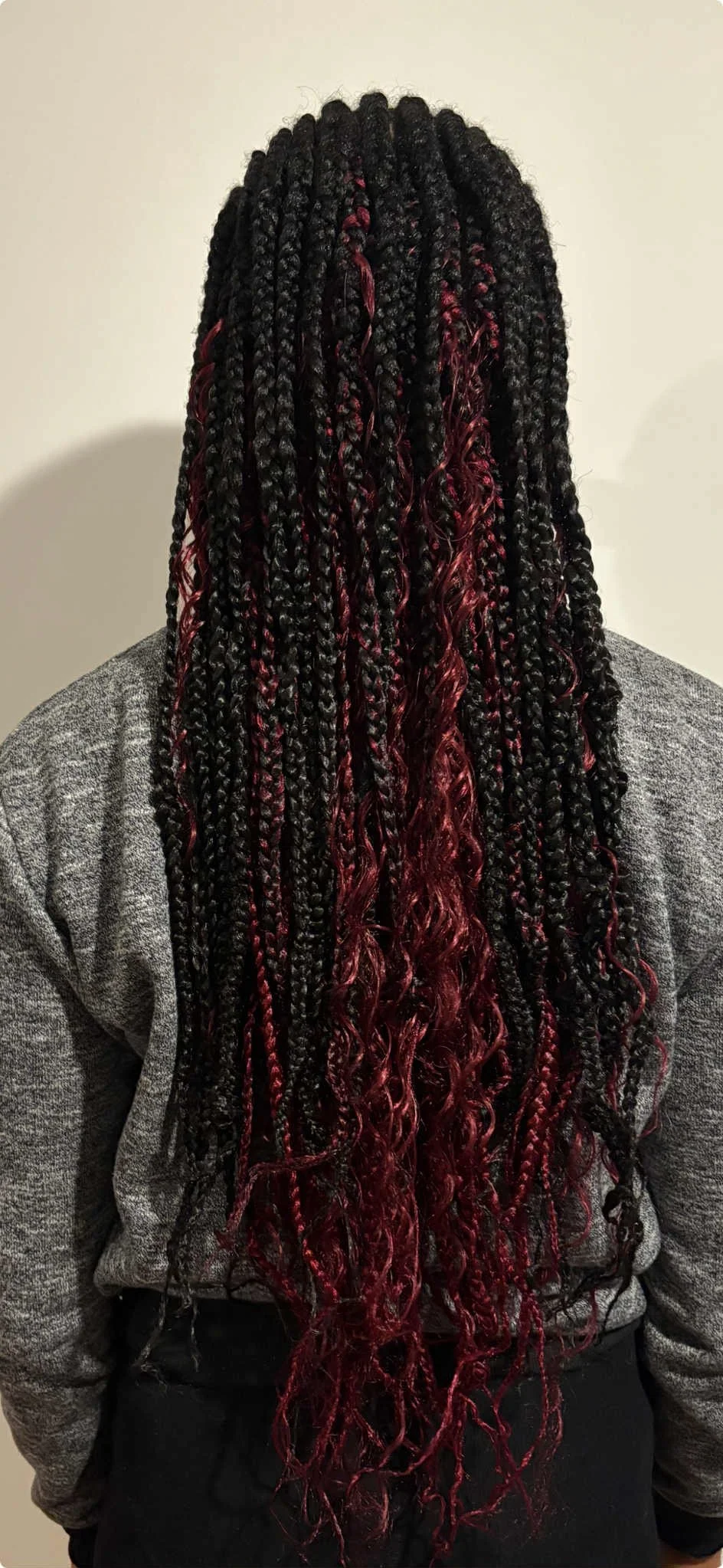 Back view of a woman with long black box braids with red accents and curls at the ends, wearing a gray sweater.