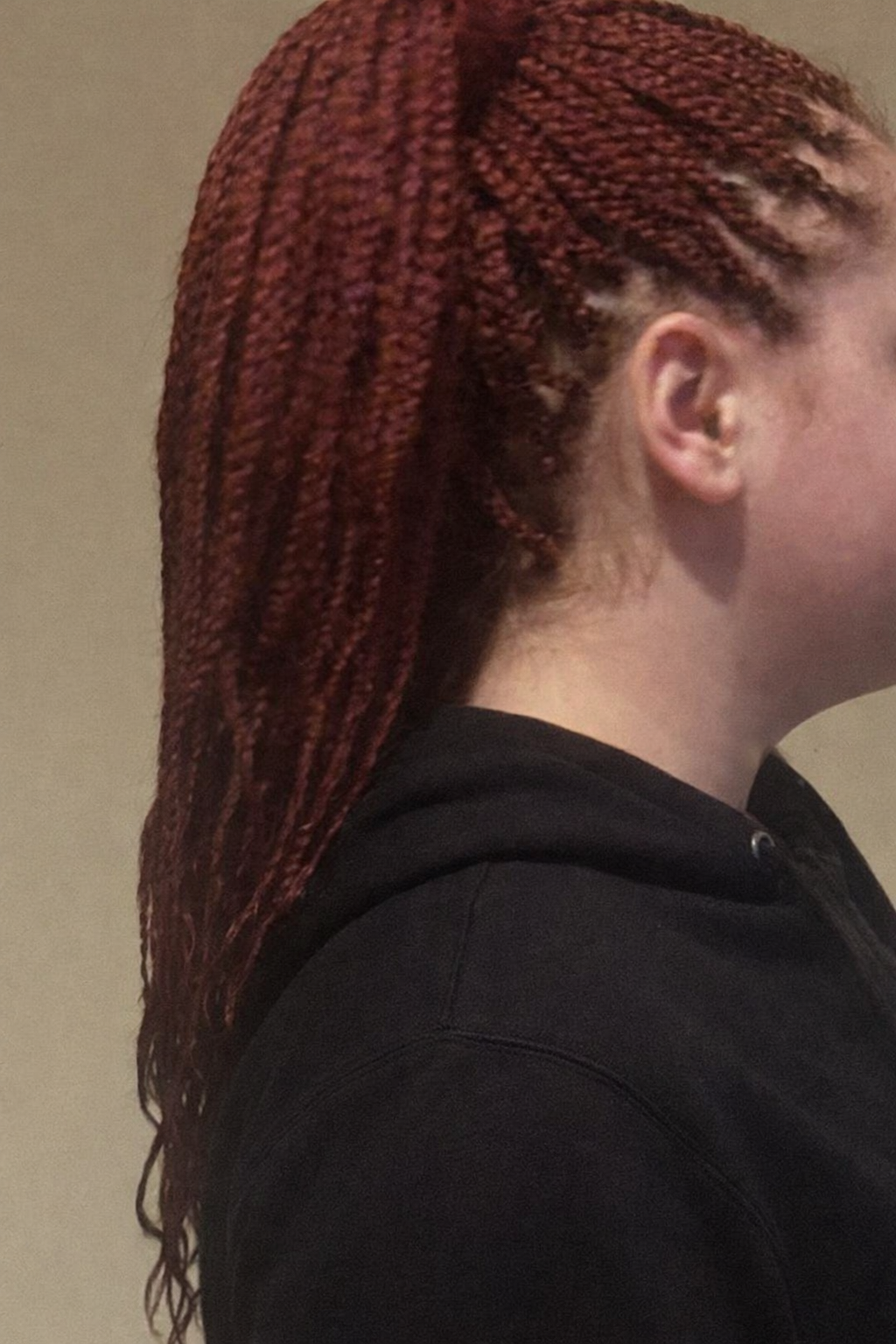 A woman with long, red, twisted braids, wearing a black hoodie, seen from the side, against a plain background.