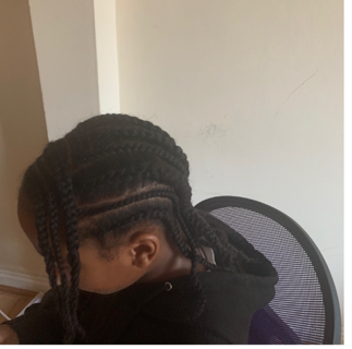 A person with braided hair sitting in a black office chair, looking down, with a plain white wall in the background.