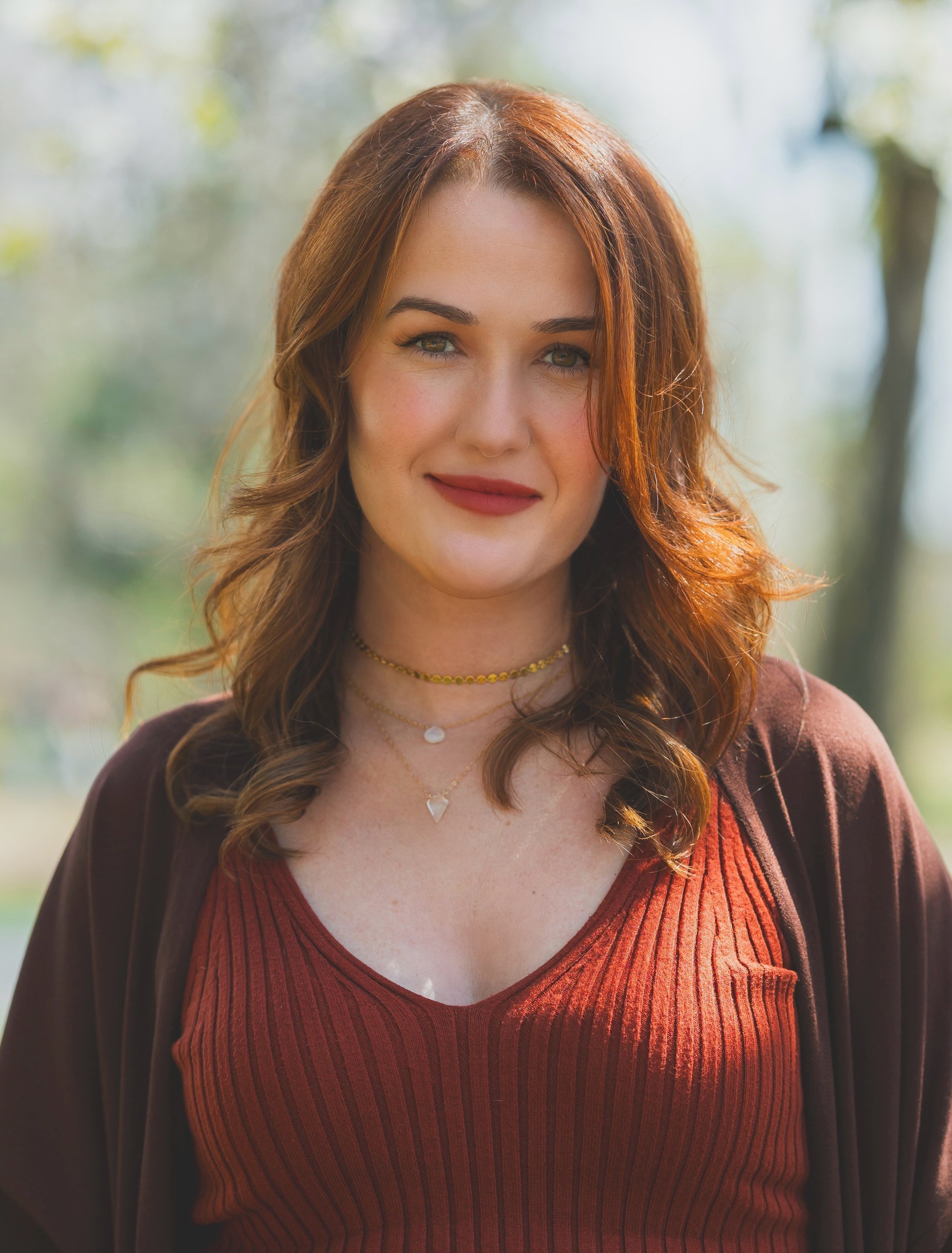 A woman with red hair wearing a brown cardigan over a red ribbed top, standing outdoors with a blurred natural background.