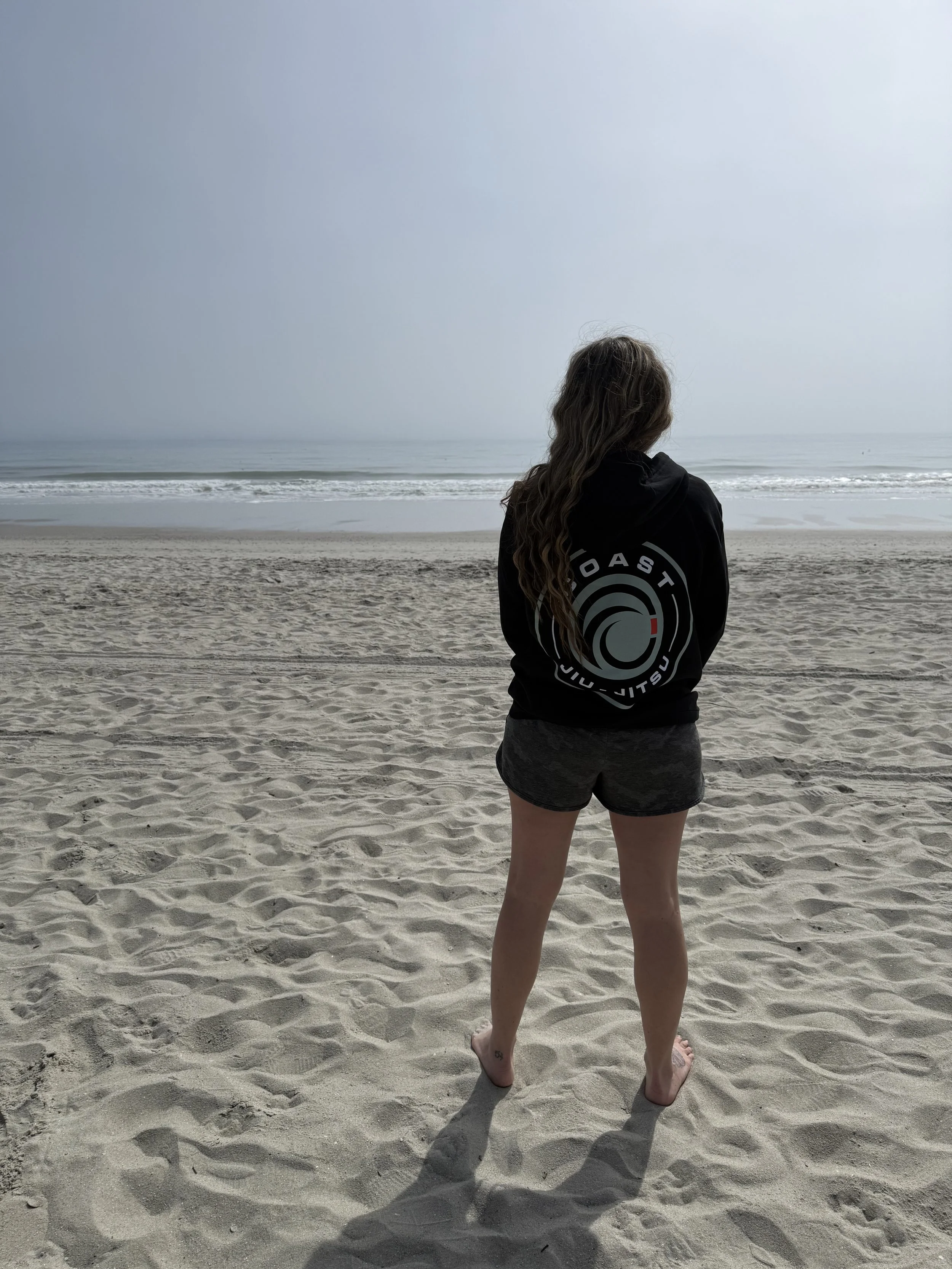 Person with long hair standing on a sandy beach, facing the ocean, wearing a black hoodie with a circular logo and dark shorts.
