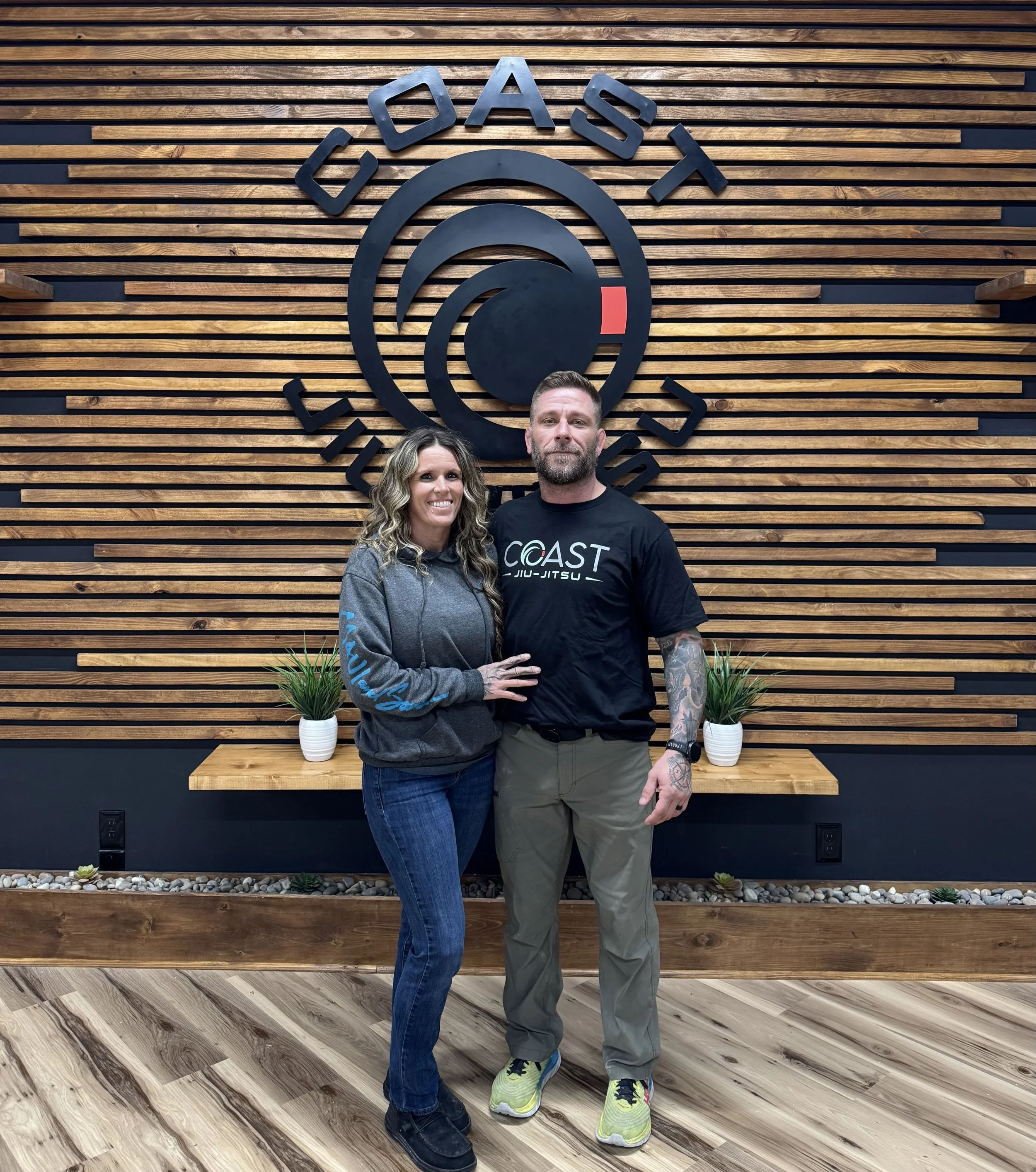 A man and woman standing in front of a wood-paneled wall with a large logo that reads 'COAST' and 'BJJ' with the words 'COAST' and 'BJJ-JITSU'. The woman is wearing a gray hoodie and jeans, and the man is wearing a black 'COAST BJJ-JITSU' t-shirt and khaki pants.