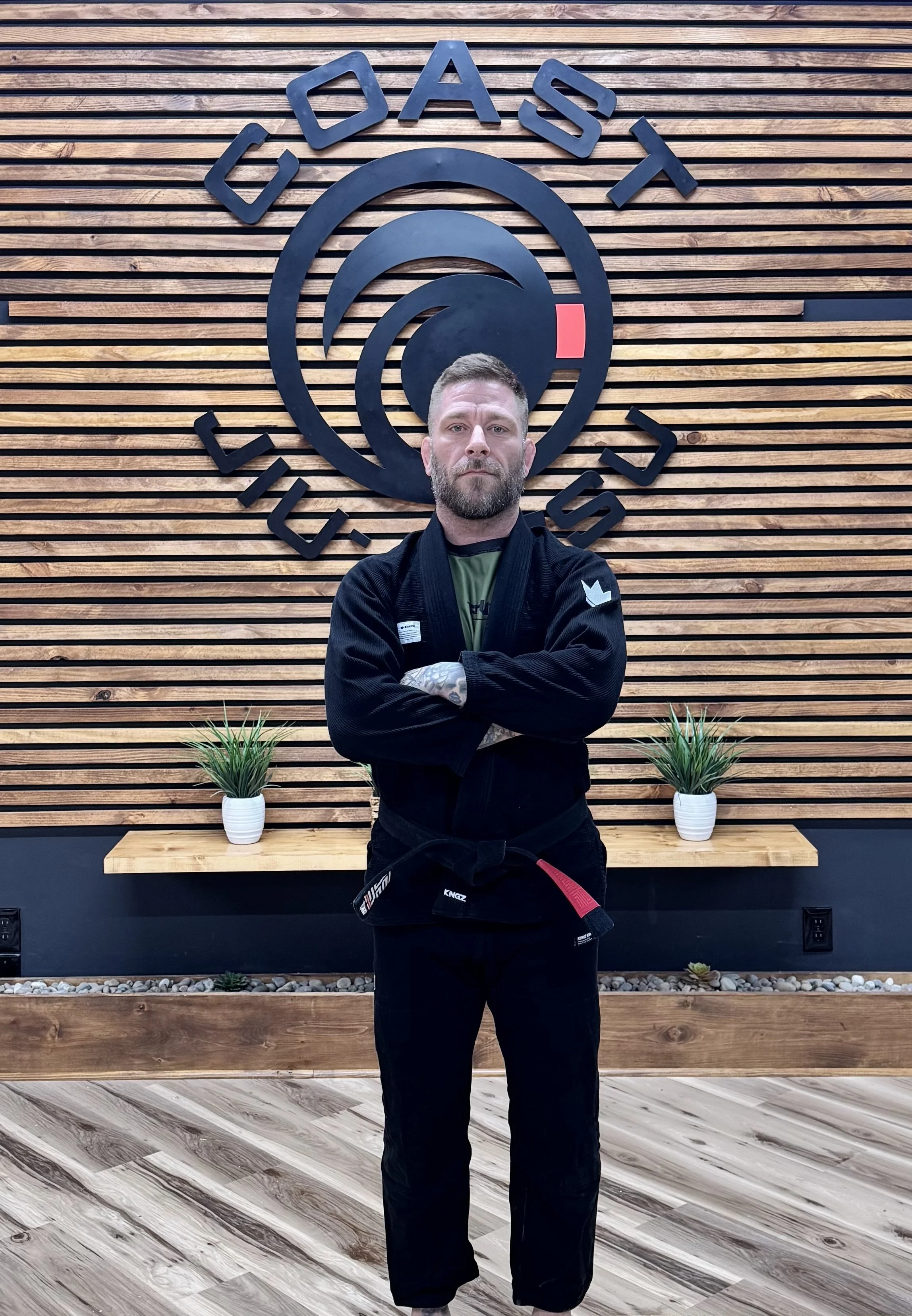 A man stands with arms crossed in a martial arts gym, wearing a black gi with a red belt. Behind him is a wooden wall with the words 'Coast Jiu-Jitsu' and a circular logo featuring a stylized wave design.