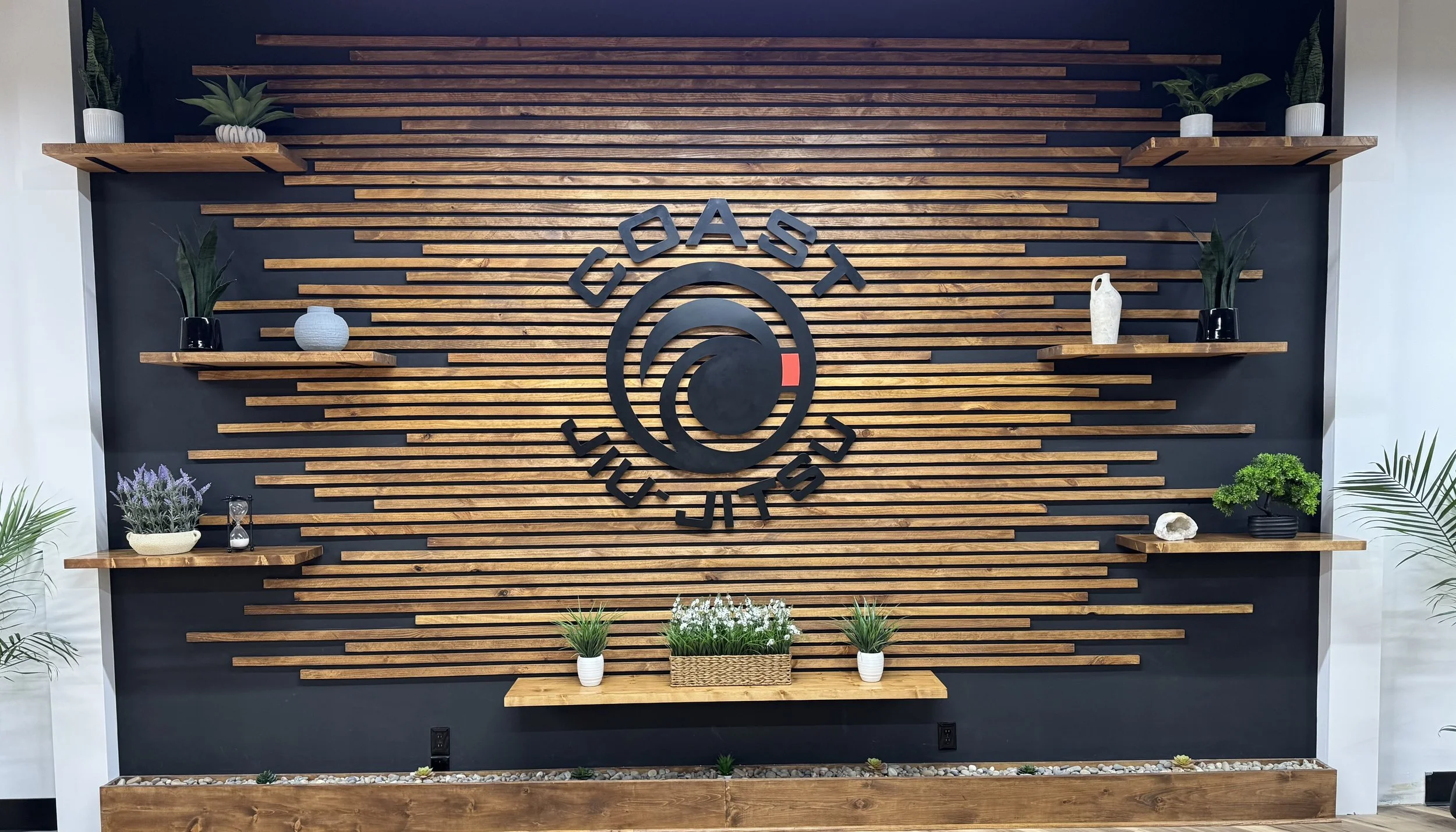 Decorative wall with wooden slats and shelves, displaying plants and ornaments, featuring the logo for 'Coast Jiu-Jitsu'