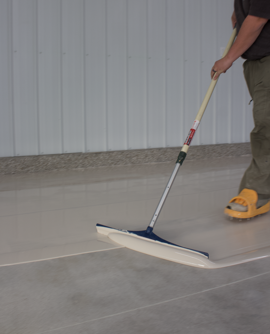 Person using a push broom to sweep a concrete floor in an indoor space.