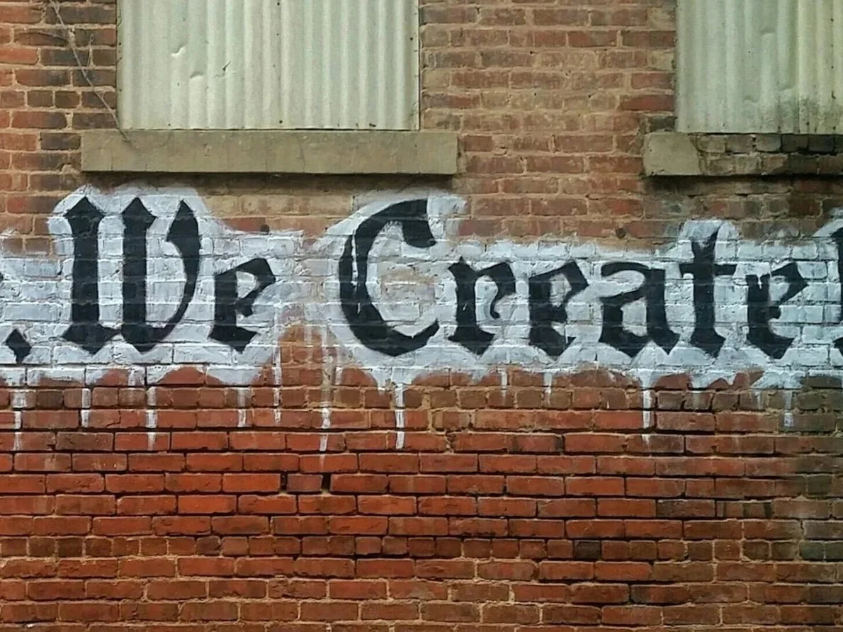 Street art graffiti on brick wall that says "De Creat" in black Old English style font with white outline.