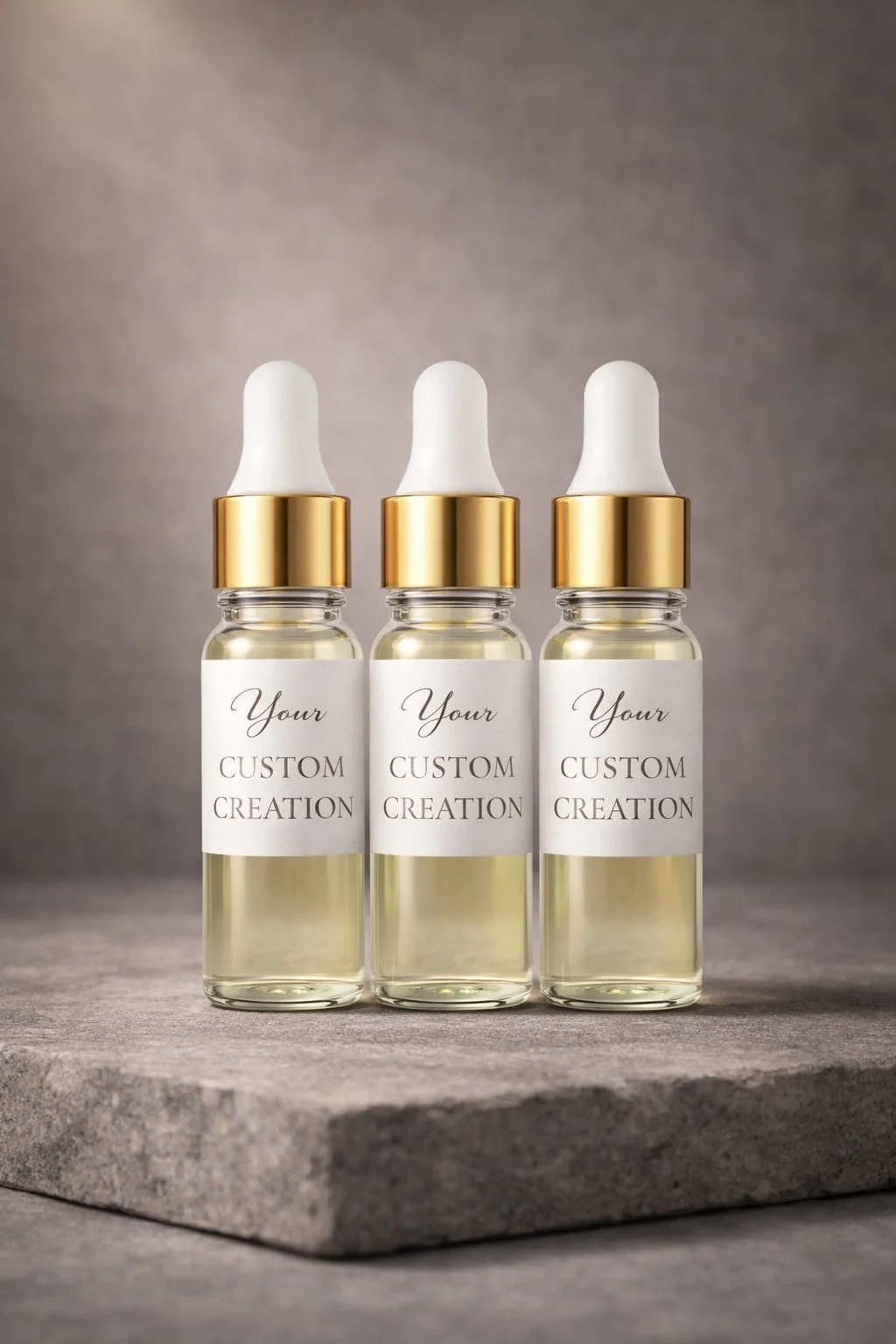 Three glass bottles with white dropper caps and gold collars, labeled 'Your Custom Creation,' are displayed on a stone surface against a gray background.
