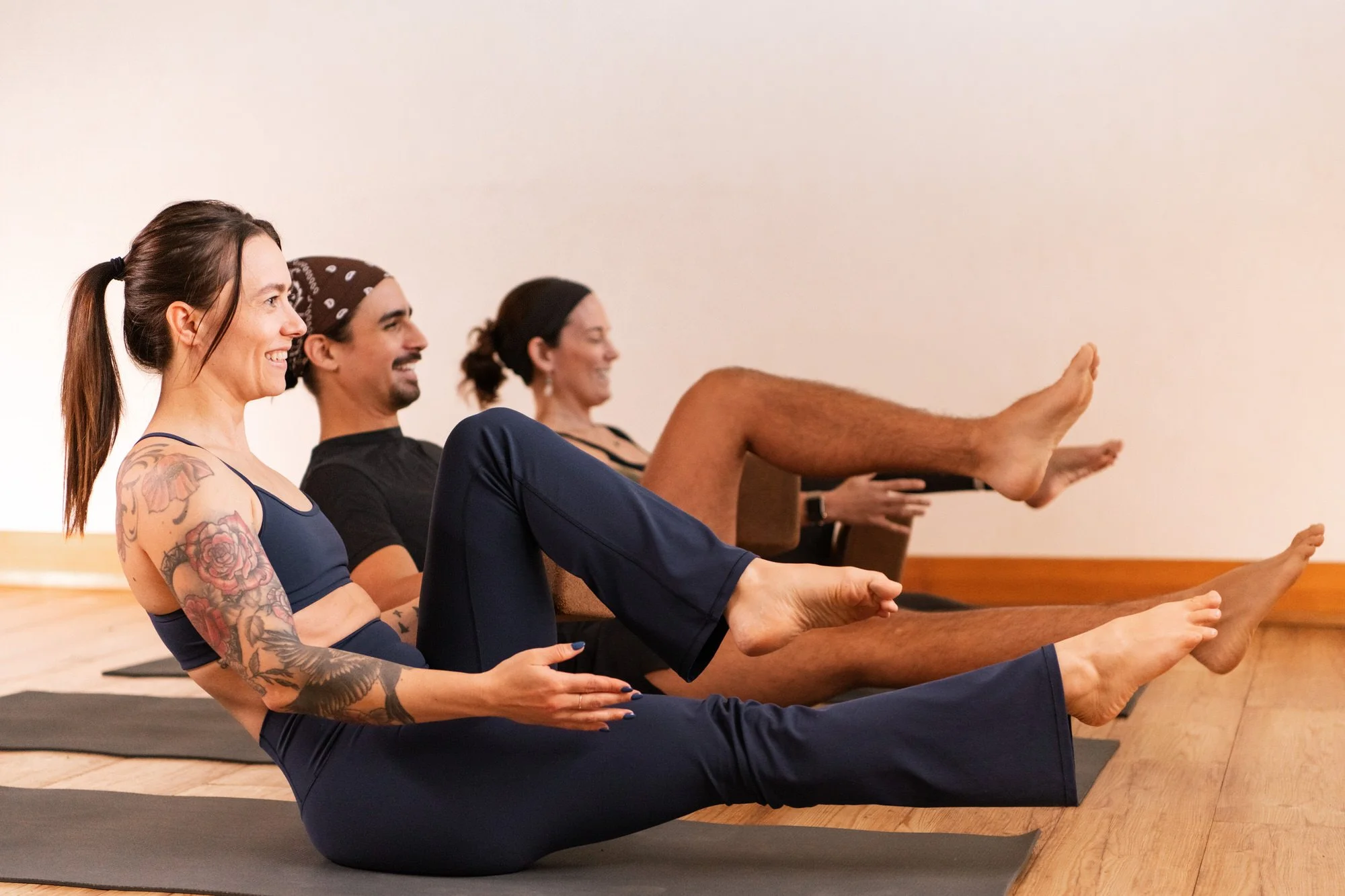 Four people participating in a Pilates class at Wild Reed Yoga in Portland, sitting on yoga mats with legs extended and balancing on their sit bones, smiling and focusing on their posture.