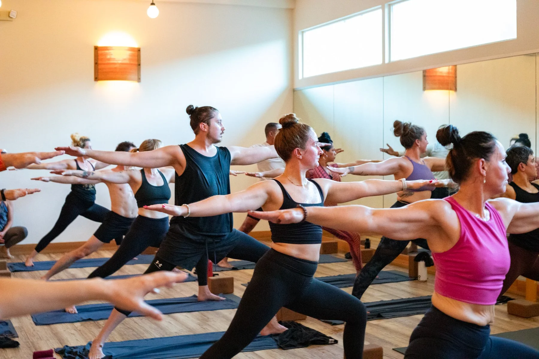 Group of people participating in a yoga class at Wild Reed Yoga in Portland, practicing Warrior pose in a well-lit studio with wooden floors and large windows.