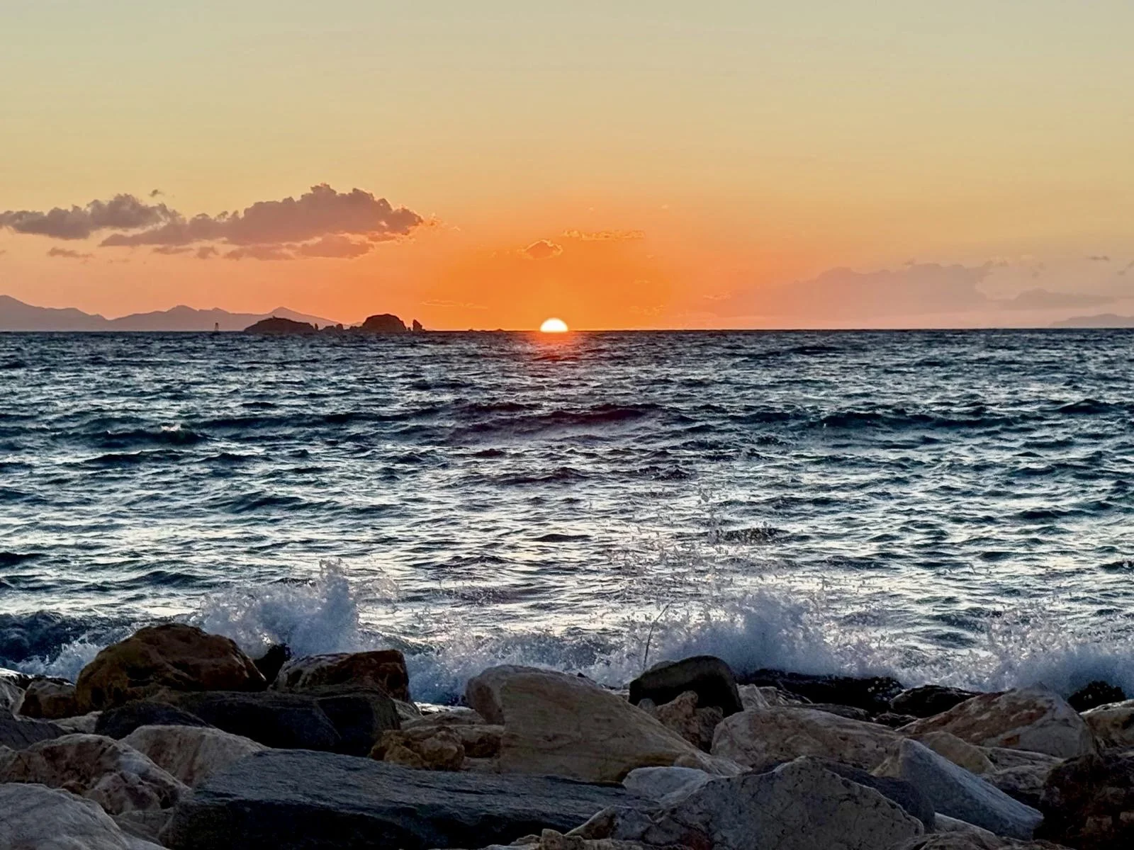 Sunset over the ocean with rocks in the foreground and small waves crashing, distant islands on the horizon.