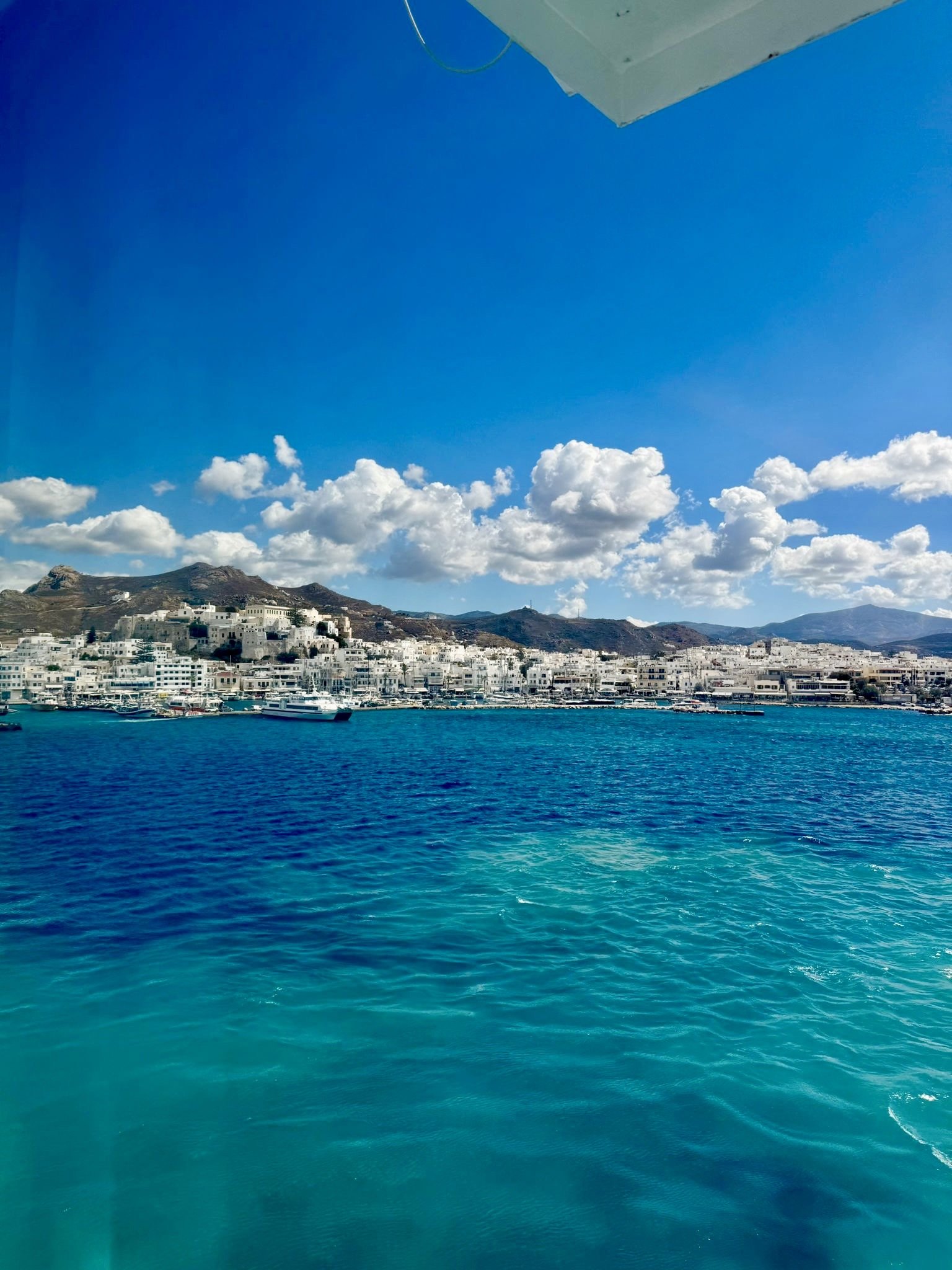 View of a coastal town with white buildings on a hillside, blue water in the foreground, and a partly cloudy sky.