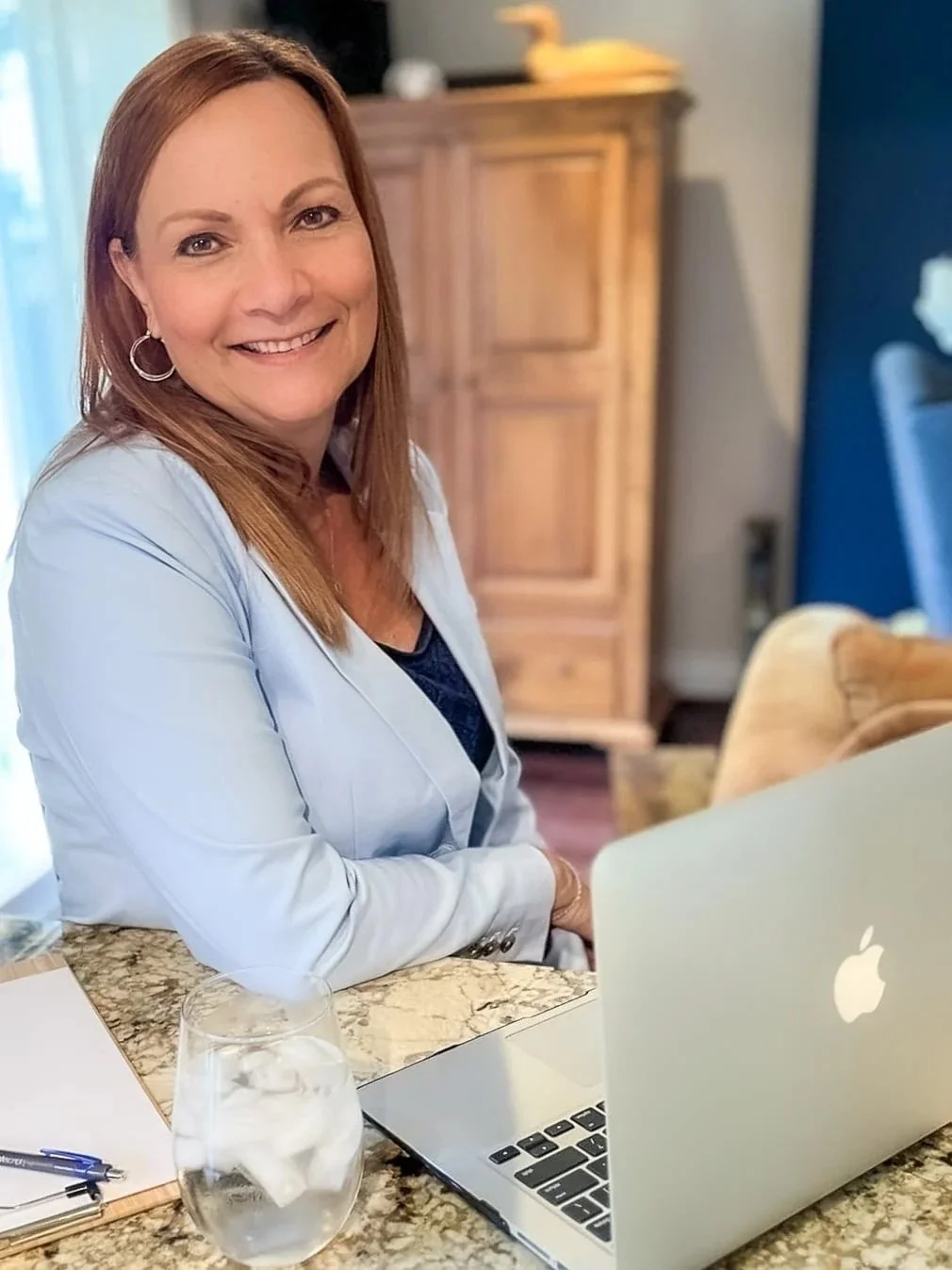 A woman with shoulder-length reddish-brown hair smiling at a camera, sitting at a kitchen counter with a silver Apple laptop in front of her, a glass of water, a notepad, and a pen. Behind her is a wooden cabinet and a blue wall.