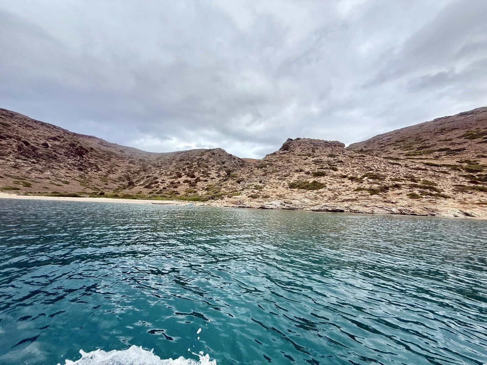 View of a rocky coastline with hills and sparse vegetation, under a cloudy sky, adjacent to a body of clear blue-green water.