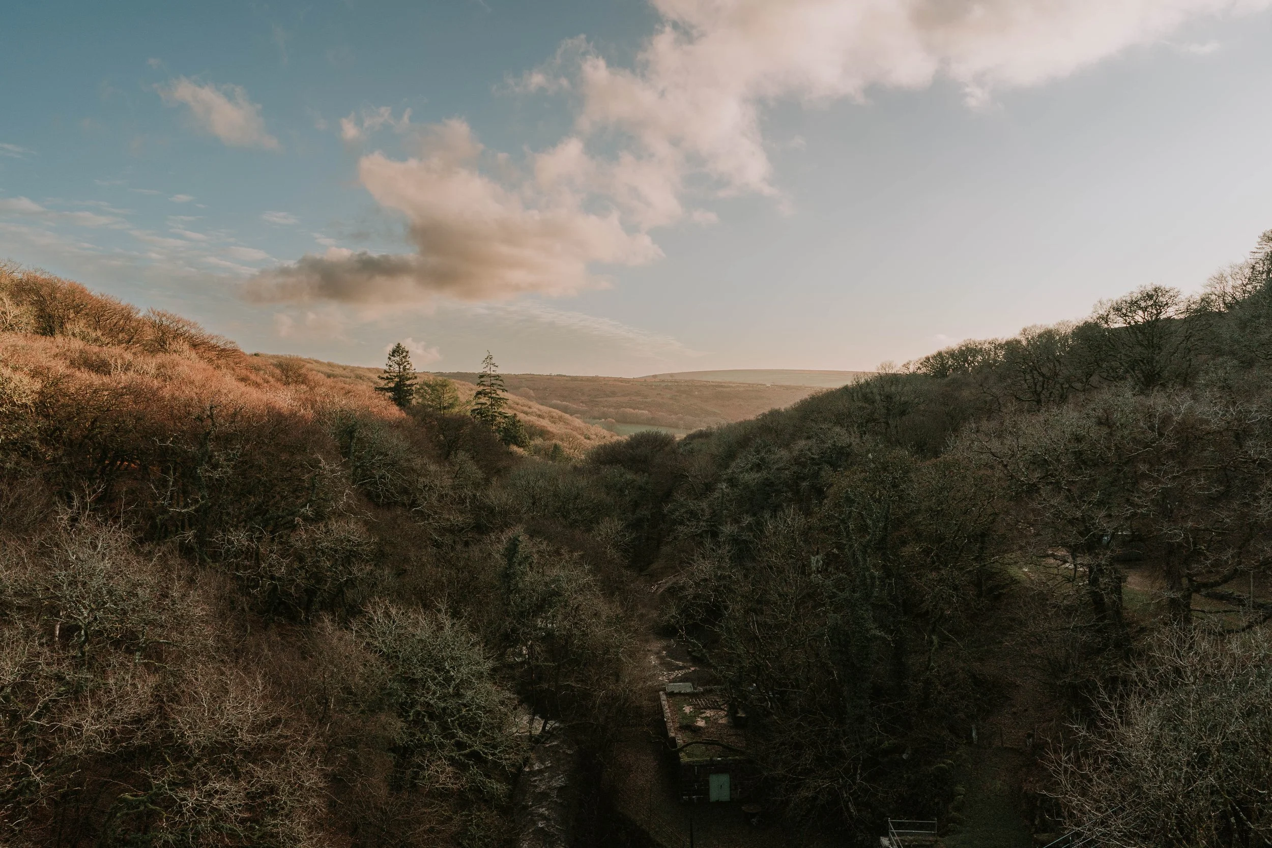 Scenic view of a valley with leafless trees and rolling hills under a partly cloudy sky at sunset or sunrise.