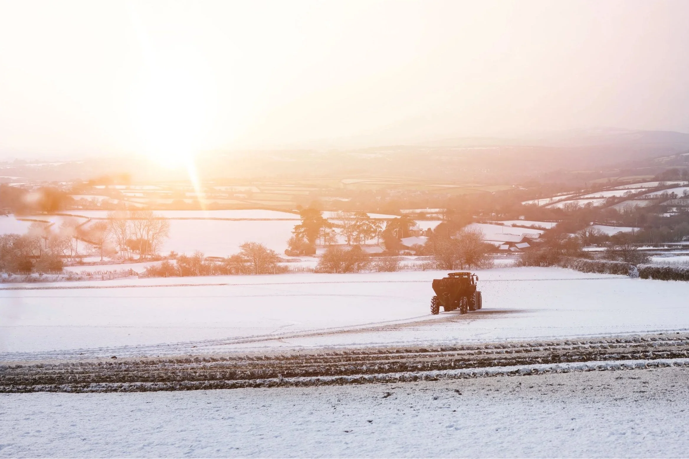 A tractor working in a snow-covered field at sunset or sunrise, with a rural landscape and hills in the background. Taken at Dowsland on Dartmoor just before sunset 