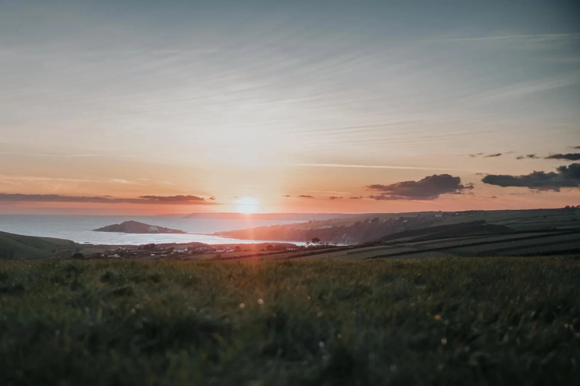 Sunset over a coastal landscape with rolling green fields, a small island, and a bay, featuring scattered clouds and a colorful sky.