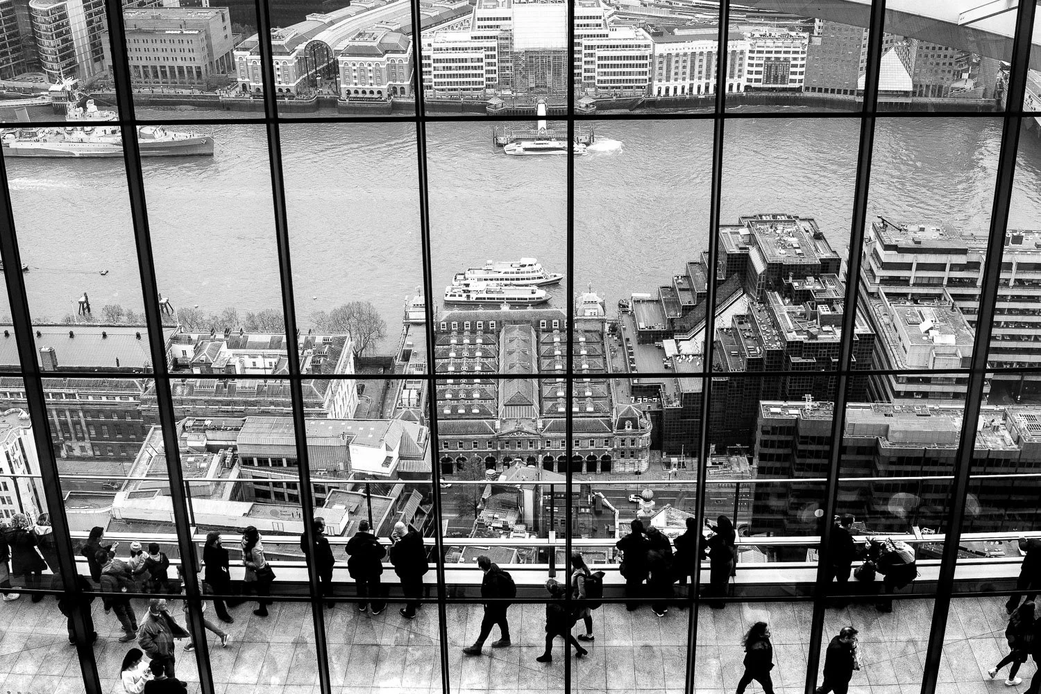 View from inside a tall building showing a cityscape with a river, boats, and surrounding buildings. People are standing on a balcony or observation deck looking out at the view. Taken at the Sky Garden in the city of London 