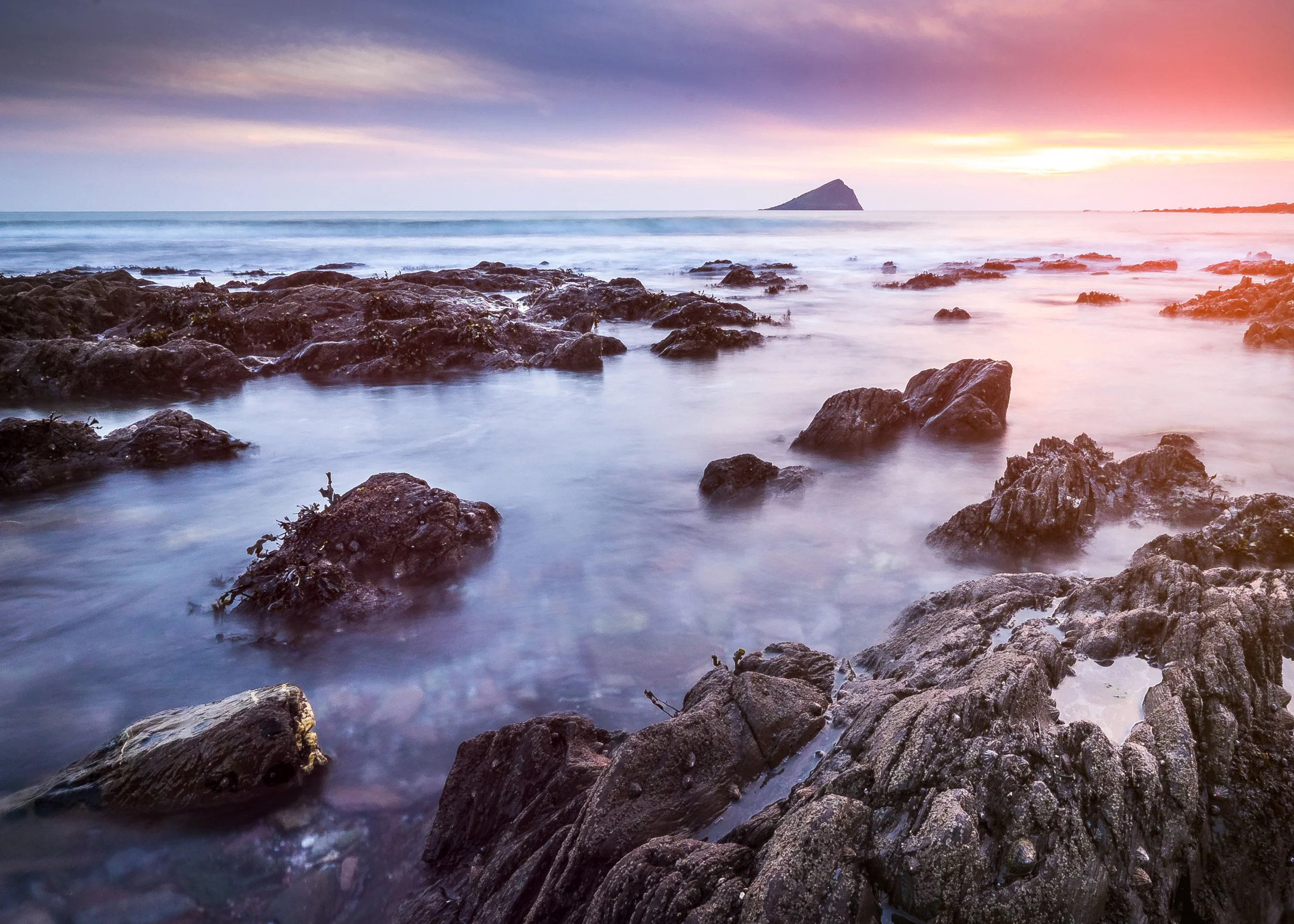 A rocky shoreline at sunset with waves softly crashing against rocks, a distant mountain or island on the horizon, and pink, purple, and orange hues in the sky. Taken at Wembury beach near plymouth at sunset 