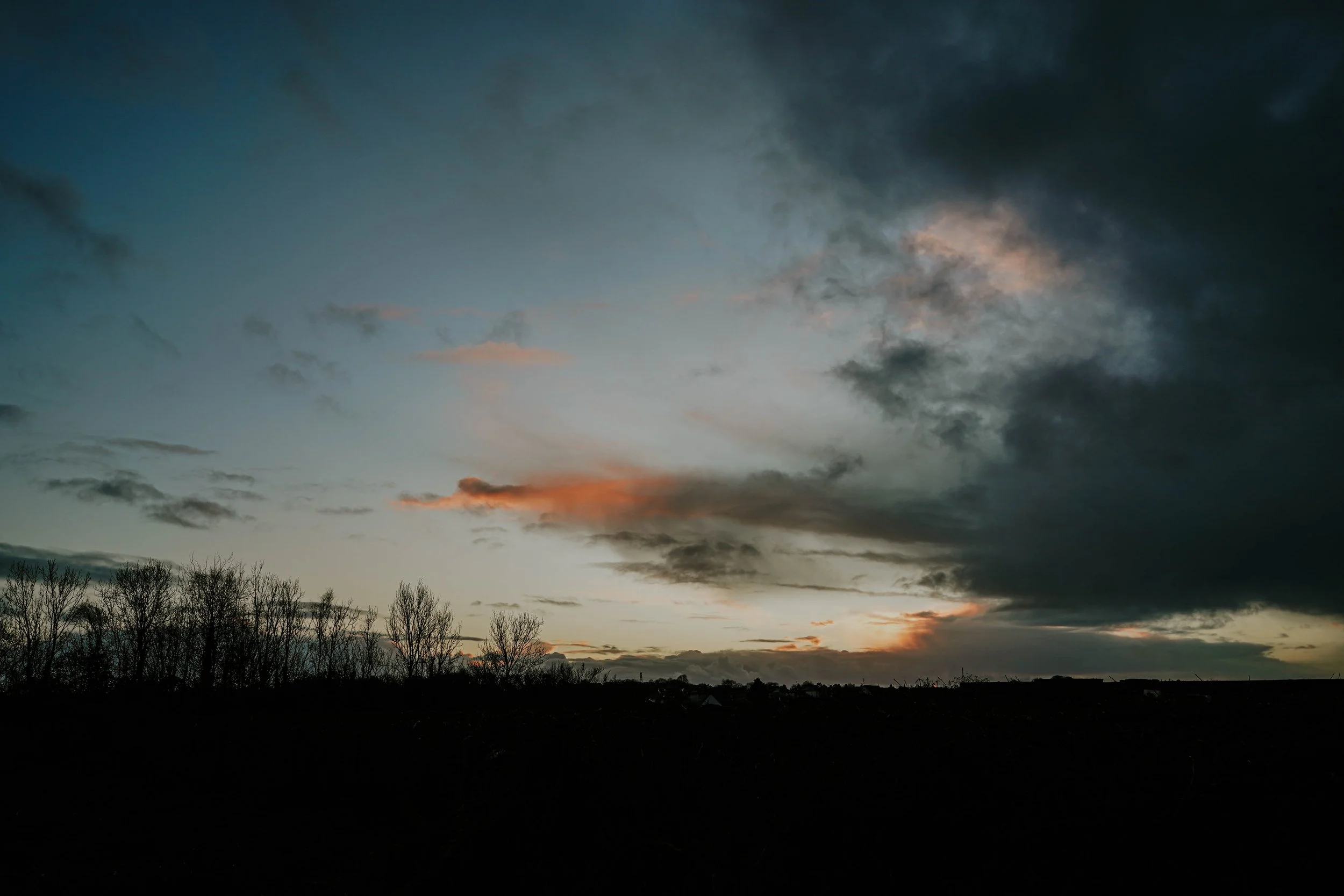 Cloudy sunset sky with dark and light clouds, silhouetted trees and landscape below.