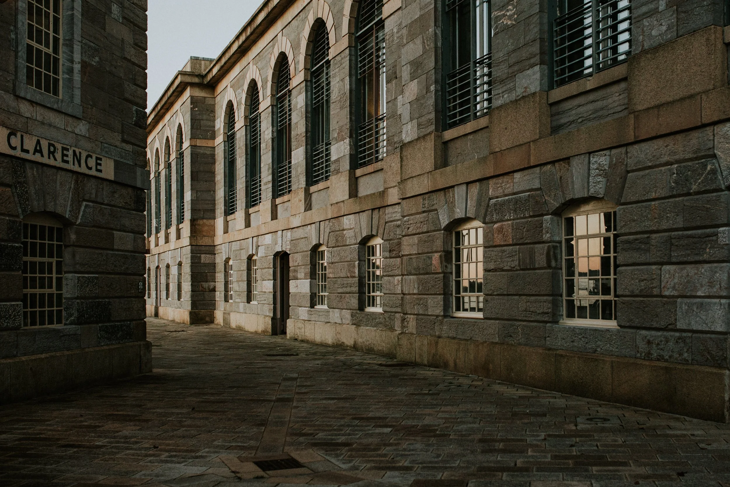 A historic stone building with arched windows and a sign that reads 'CLARENCE' on the corner, illuminated by soft evening light. Taken at the Royal William Yard in Plymouth 