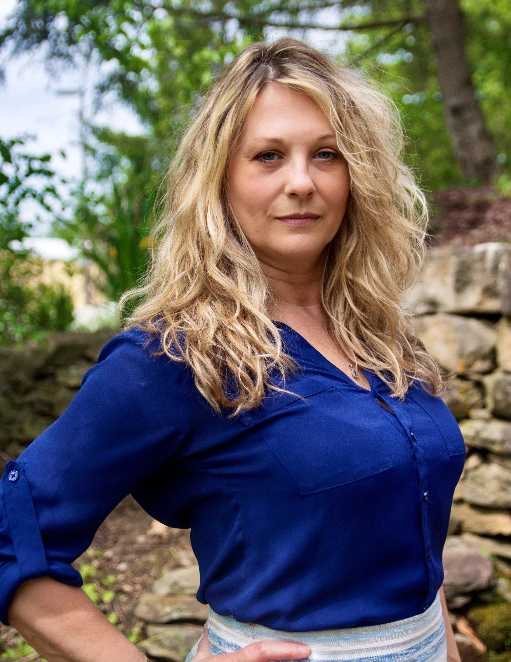 Toni Antonucci is a woman with blonde hair in loose waves wearing a blue shirt standing outdoors in front of trees and rocks.
