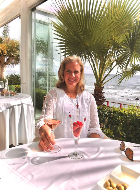 Gabrielle England artist sitting at a table with a pink cocktail and a cherry dessert, smiling, with beach and ocean view in the background.