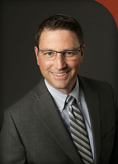 Portrait of a smiling man wearing glasses, a gray suit, a white shirt, and a striped tie against a dark background.