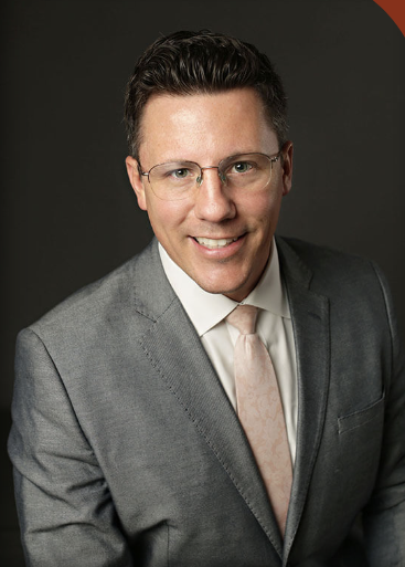 Professional headshot of a man in a gray suit, white shirt, and light pink tie, smiling against a dark background.