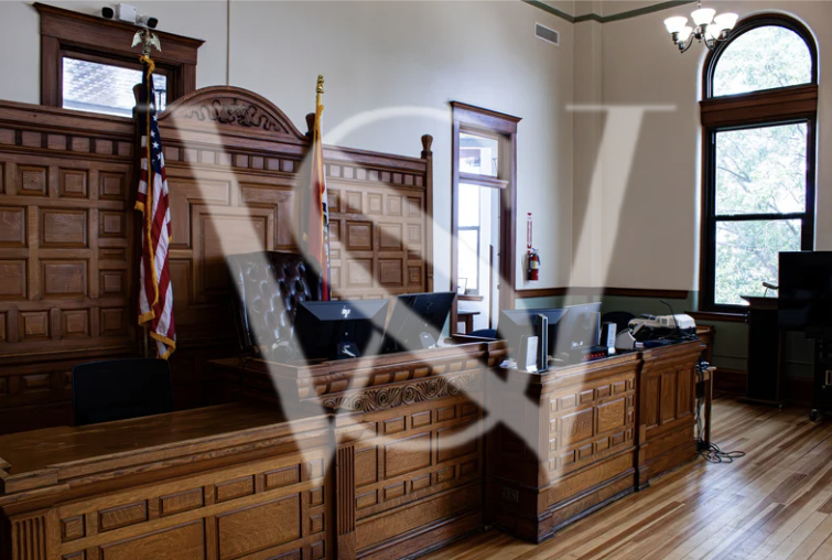 Inside a courtroom with wooden paneling, a judge's bench, and American flags, large windows, and a fire extinguisher on the wall.