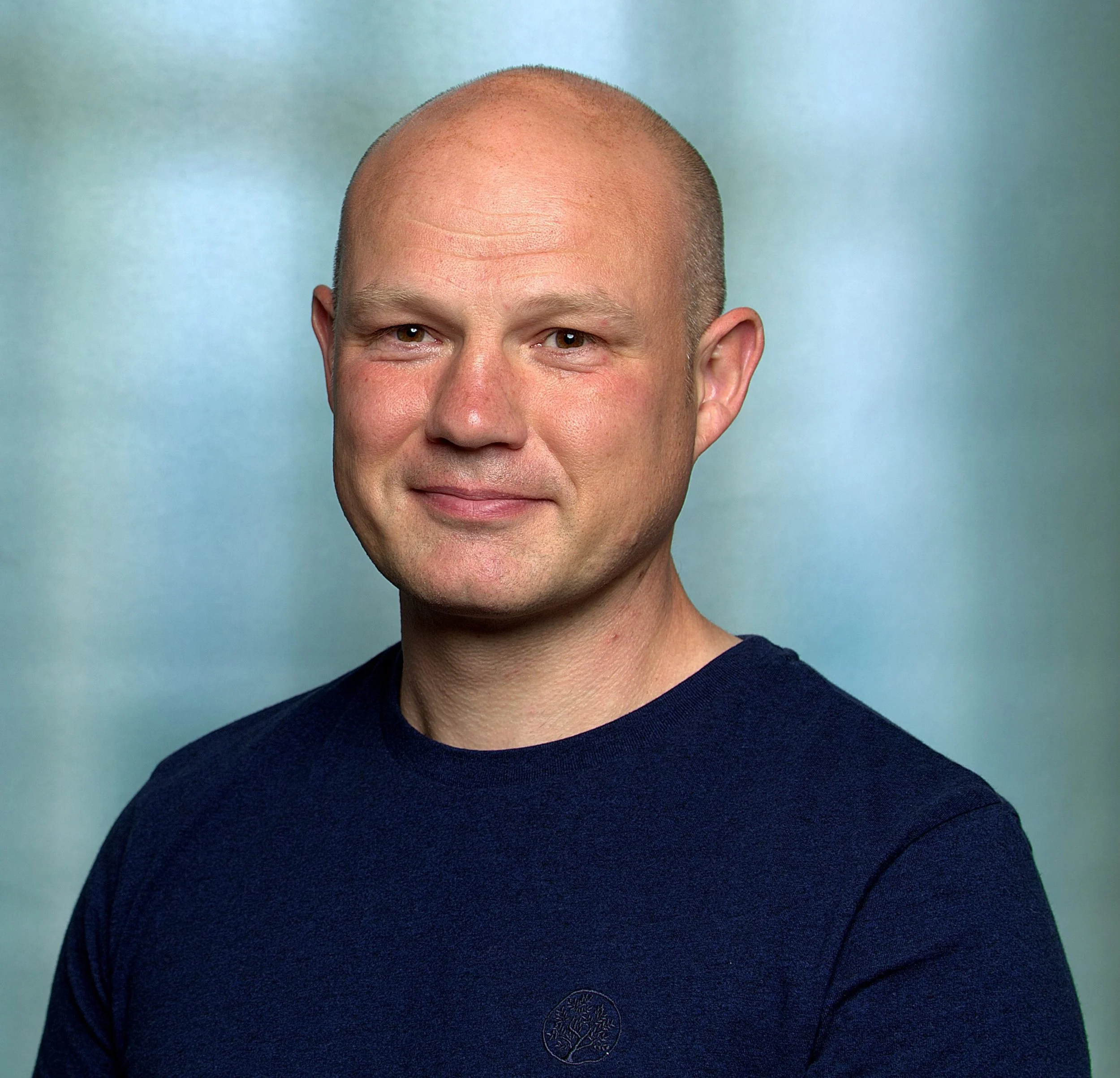 Headshot of a smiling man with a shaved head, wearing a navy shirt, against a light blue background.