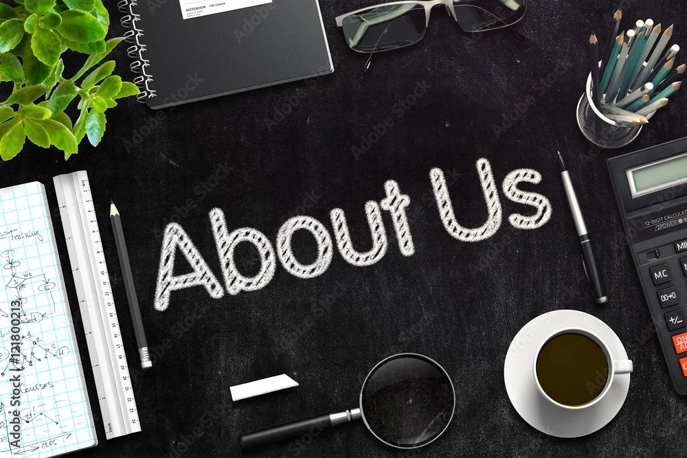 Top view of a workspace with the words "About Us" written in chalk on a blackboard. The workspace includes notebooks, glasses, sunglasses, pencils, a calculator, a cup of coffee, and various office supplies.